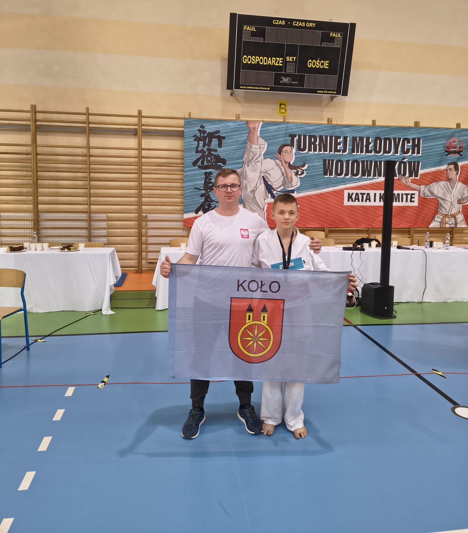 Na zdjęciu widać mężczyznę i chłopca trzymających flagę miasta Koła. Chłopiec jest ubrany w strój kimono, a na szyi ma zawieszony medal. Za nimi widać drabinki do ćwiczeń, na nich wiszący baner z napisem Turniej Młodych Wojowników.