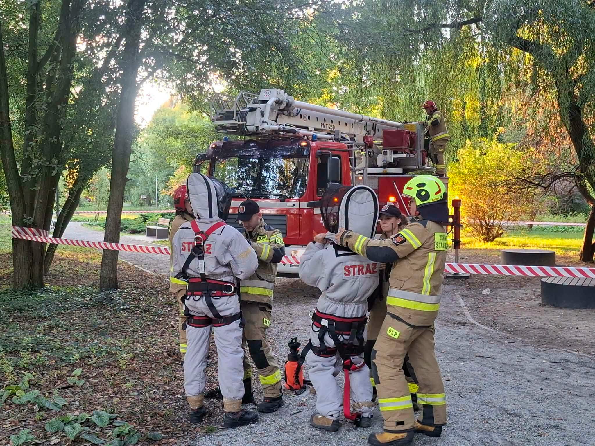 Strażacy w parku pomagają dwóm osobom założyć szare kombinezony ochronne i szelki asekuracyjne. W tle czerwony samochód pożarniczy z podnośnikiem i strażak na koszu. Teren odgrodzony biało-czerwoną taśmą.