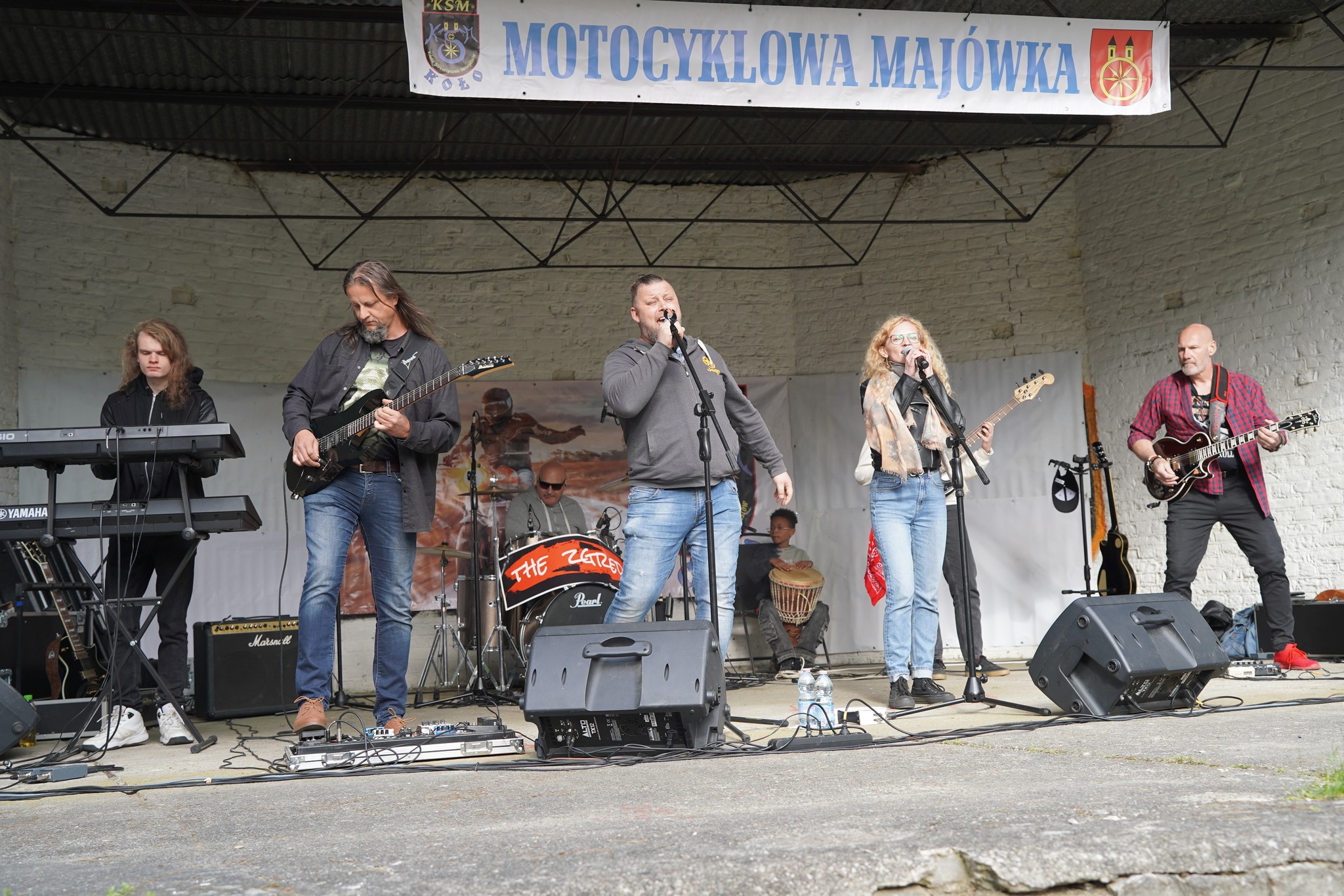 Zdjęcie przedstawia zespół muzyczny występujący na scenie podczas wydarzenia Motocyklowa Majówka – muzycy grają na żywo, a nad nimi widnieje baner imprezy.