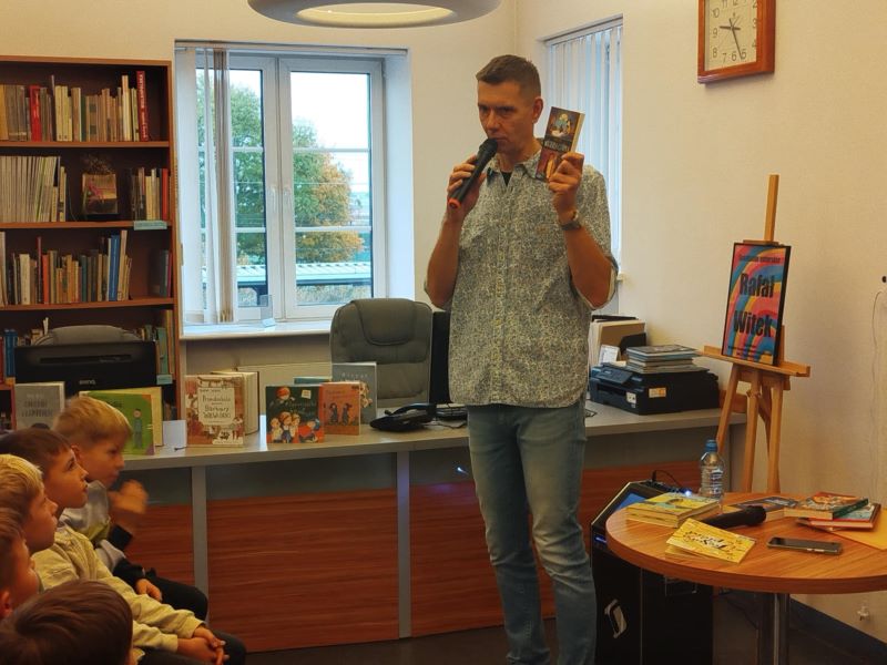 Mężczyzna z mikrofonem prowadzi spotkanie z dziećmi w bibliotece, pokazując książkę.
