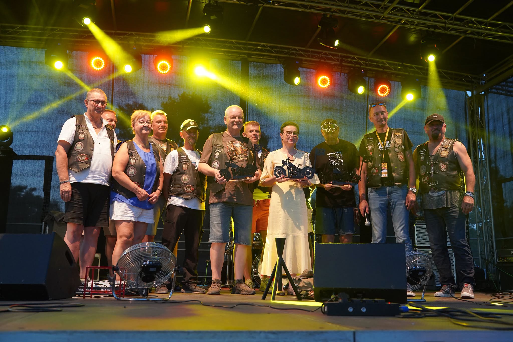 Grupa około dwunastu osób stojących na scenie podczas imprezy plenerowej. Kilka osób trzyma statuetki w kształcie motocykla. Większość uczestników ubrana w skórzane kamizelki z naszywkami. W tle kolorowe oświetlenie sceniczne.