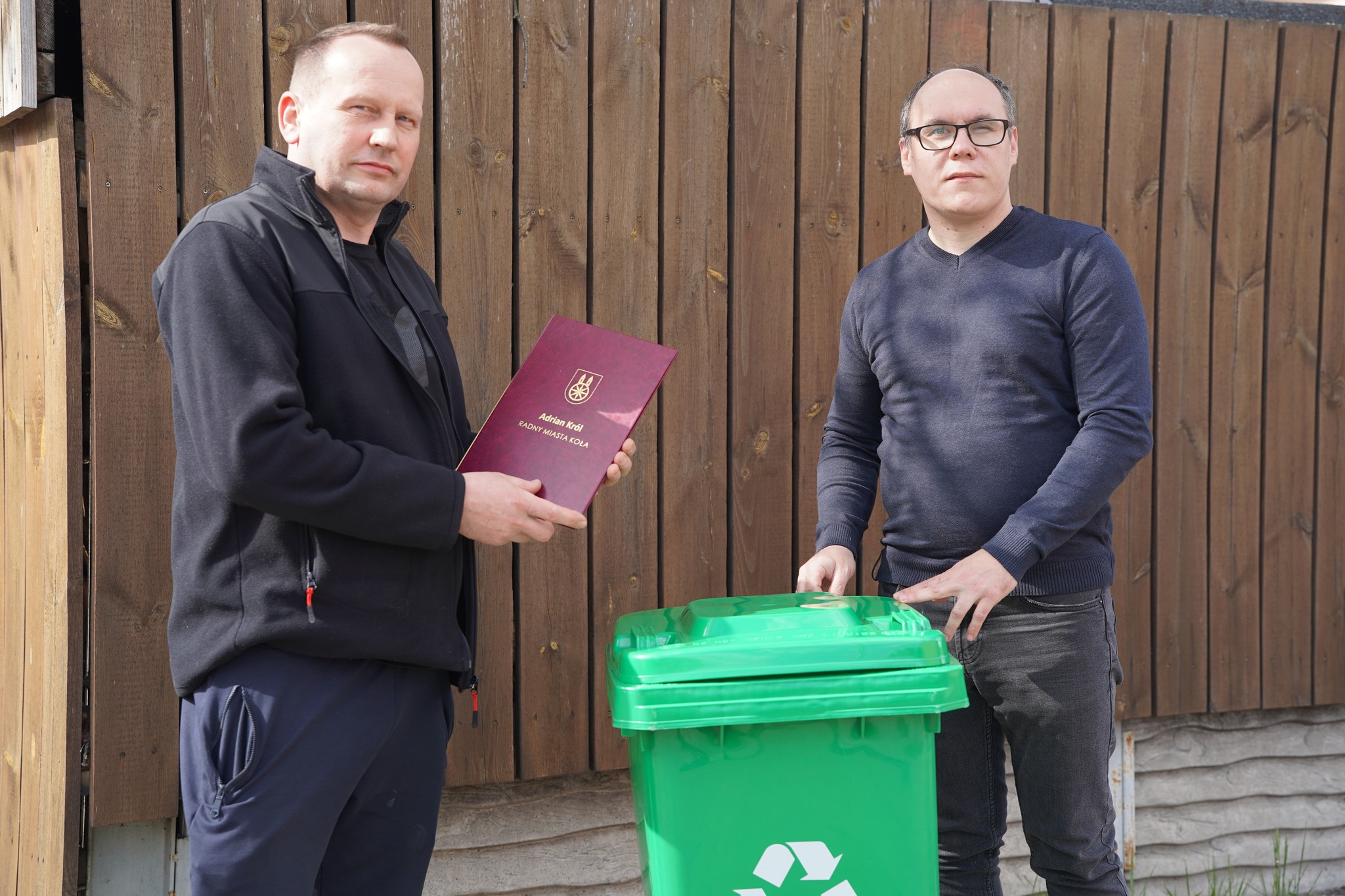 Radny Miejski Adrian Król przekazuje mieszkańcowi Koła zielony pojemnik do selektywnej zbiórki odpadów komunalnych. Mieszkaniec trzyma w dłoniach pamiątkowy dyplom, w tle widać drewniane ogrodzenie.