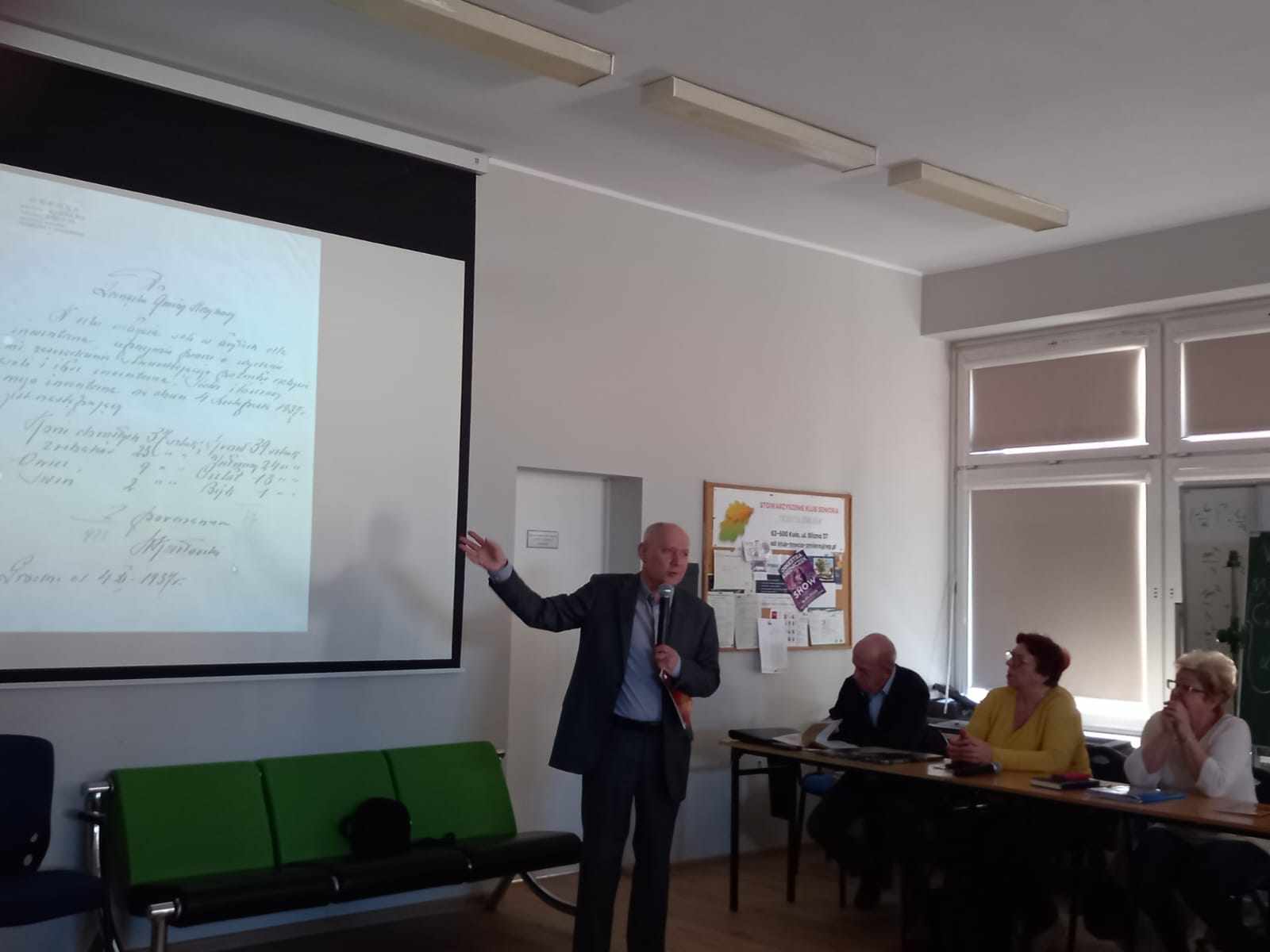 Na zdjęciu widać mężczyznę w garniturze, który prowadzi prezentację. W lewej dłoni trzyma mikrofon, prawą ręką wskazuje na wiszący na ścianie ekran, na którym wyświetlany jest slajd prezentacji. Z prawej strony siedzi kilku słuchaczy przy stole.