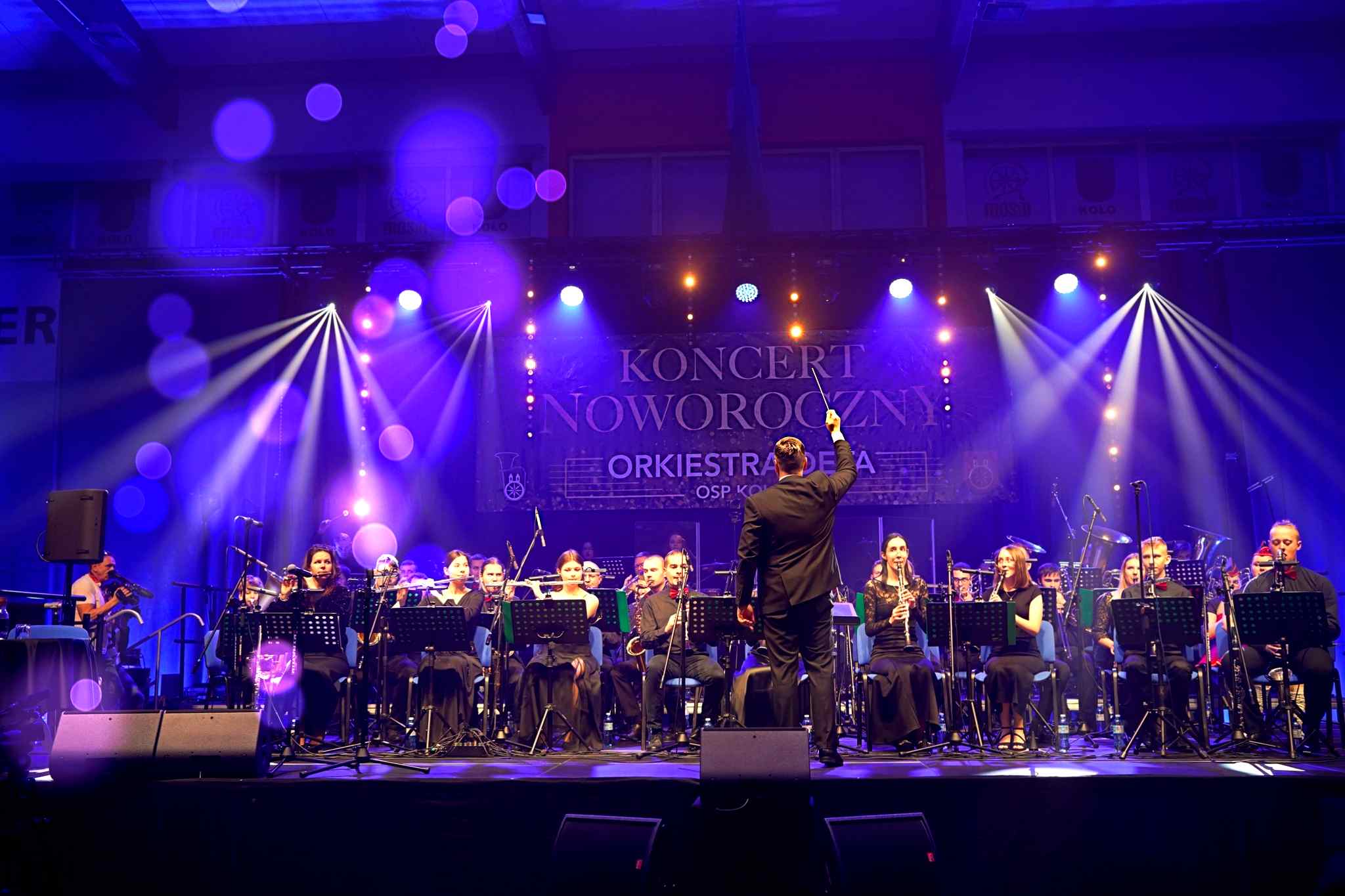Na zdjęciu widać stojącego tyłem dyrygenta z podniesioną batutą w prawej dłoni na tle orkiestry. Tło stanowi oświetlony baner z napisem KONCERT NOWOROCZNY ORKIESTRA DĘTA OSP KOŁO.