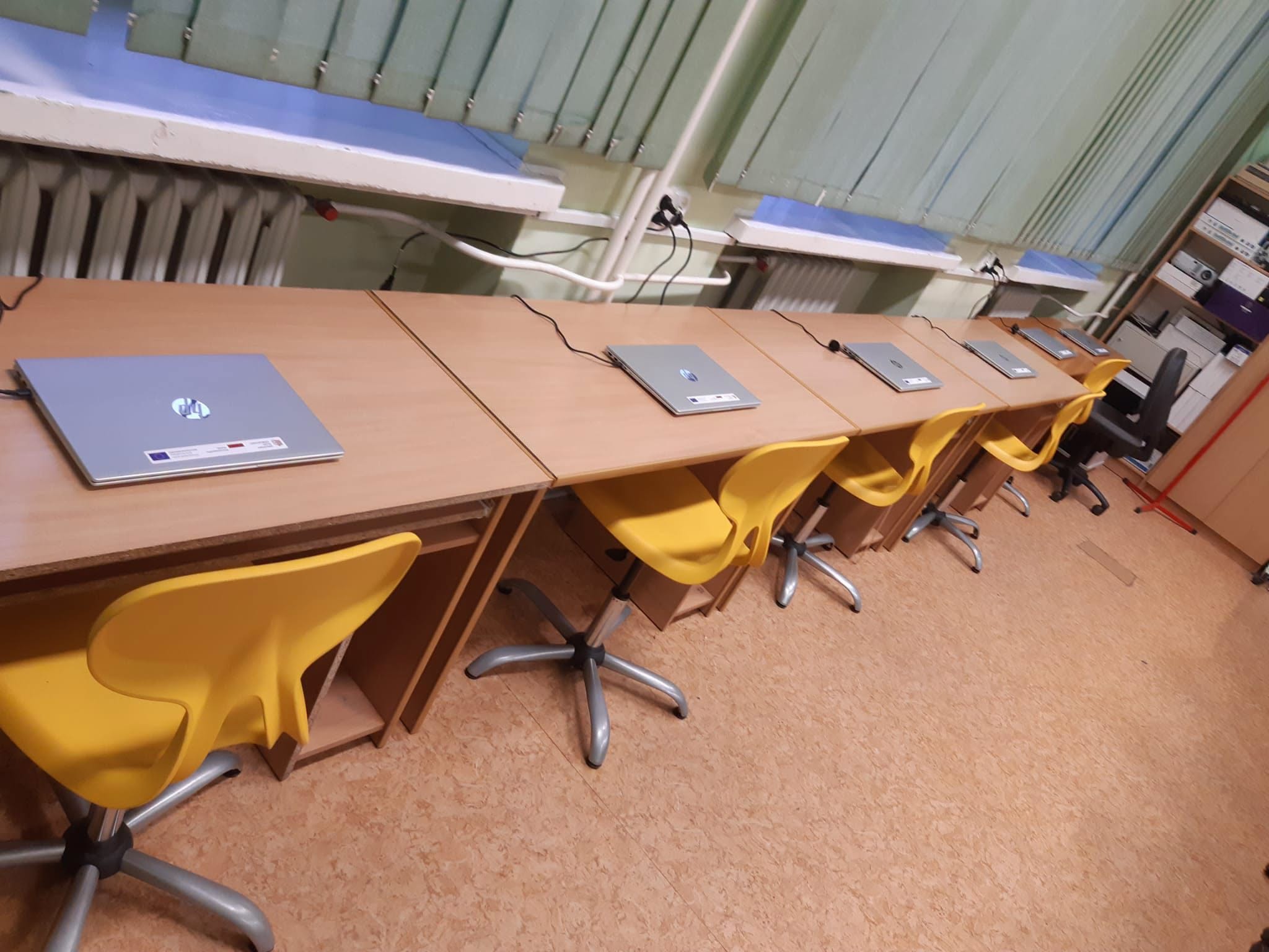 Sala lekcyjna z długim rzędem połączonych jasnych stolików ustawionych pod oknem z zieloną żaluzją pionową. Na blatach pięć zamkniętych srebrnych laptopów, przy nich żółte obrotowe krzesła na metalowych podstawach.