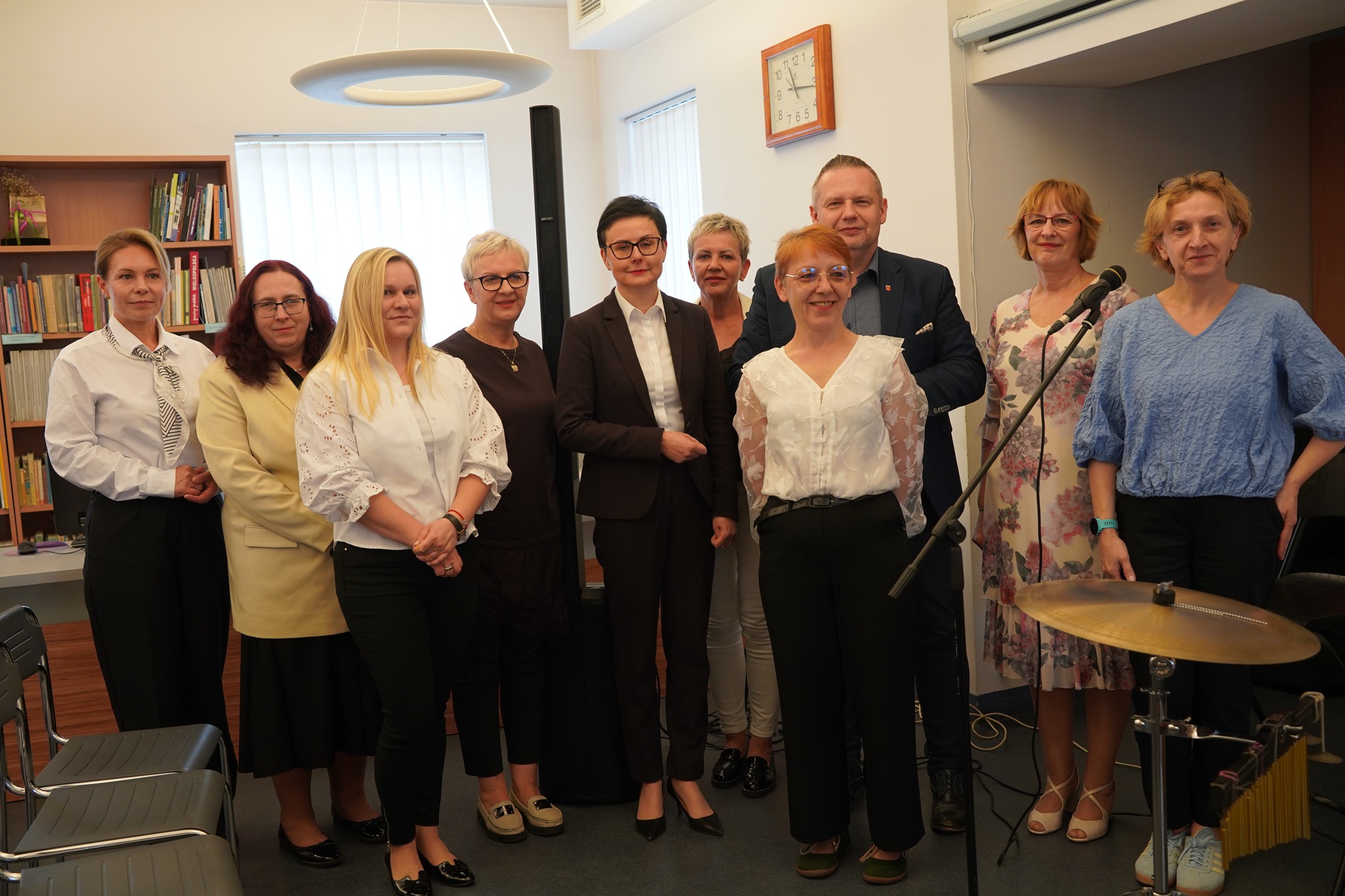 Grupa dziewięciu osób stoi w jasnym wnętrzu biblioteki. Uczestnicy pozują do wspólnego zdjęcia obok mikrofonu i instrumentów, ubrani elegancko i półformalnie.