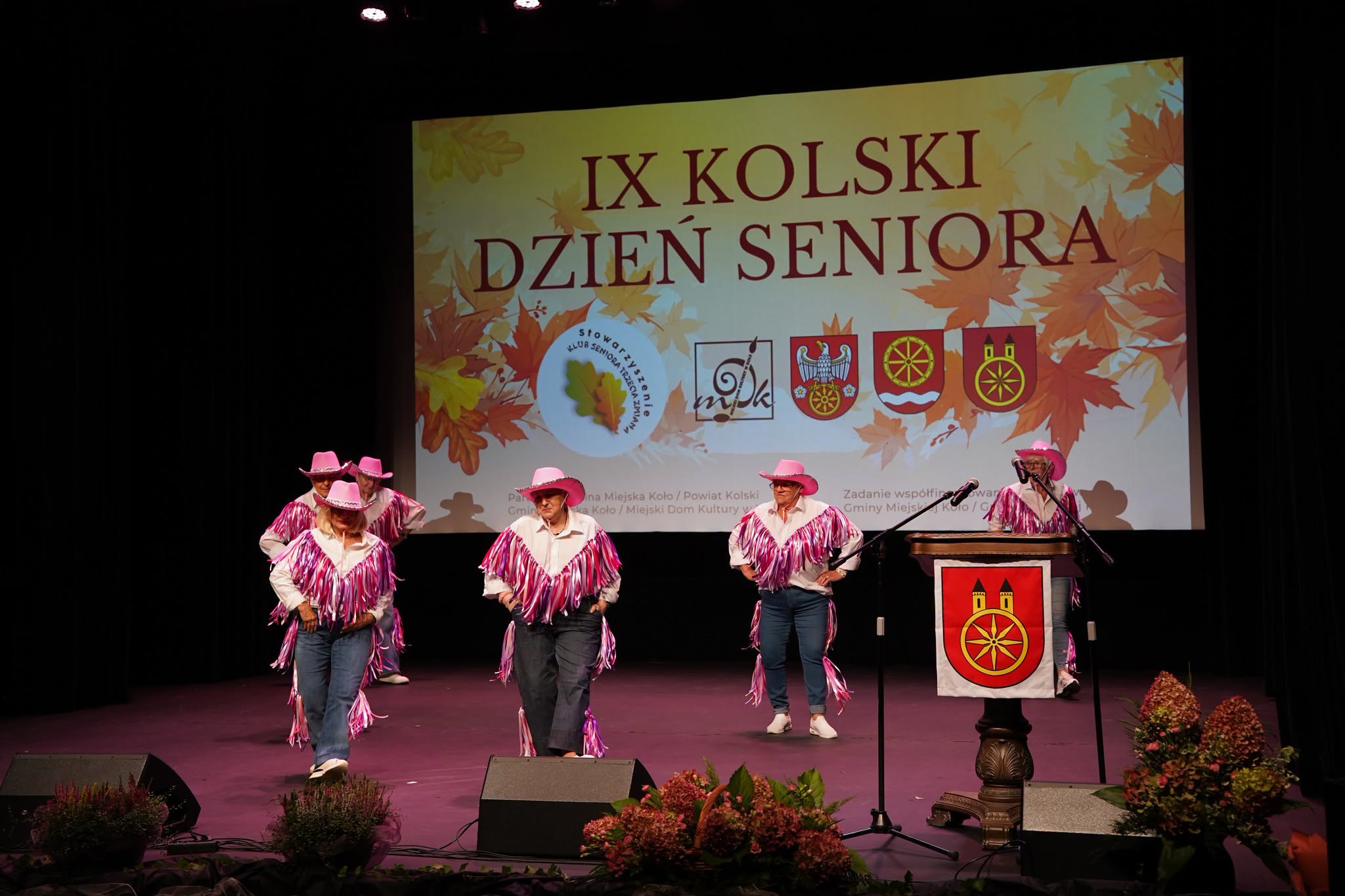 Seniorki w różowych kowbojskich kapeluszach, białych koszulach z frędzlami i dżinsach tańczą na scenie. W tle ekran z napisem IX Kolski Dzień Seniora, z prawej mównica z herbem Koła i kwiaty