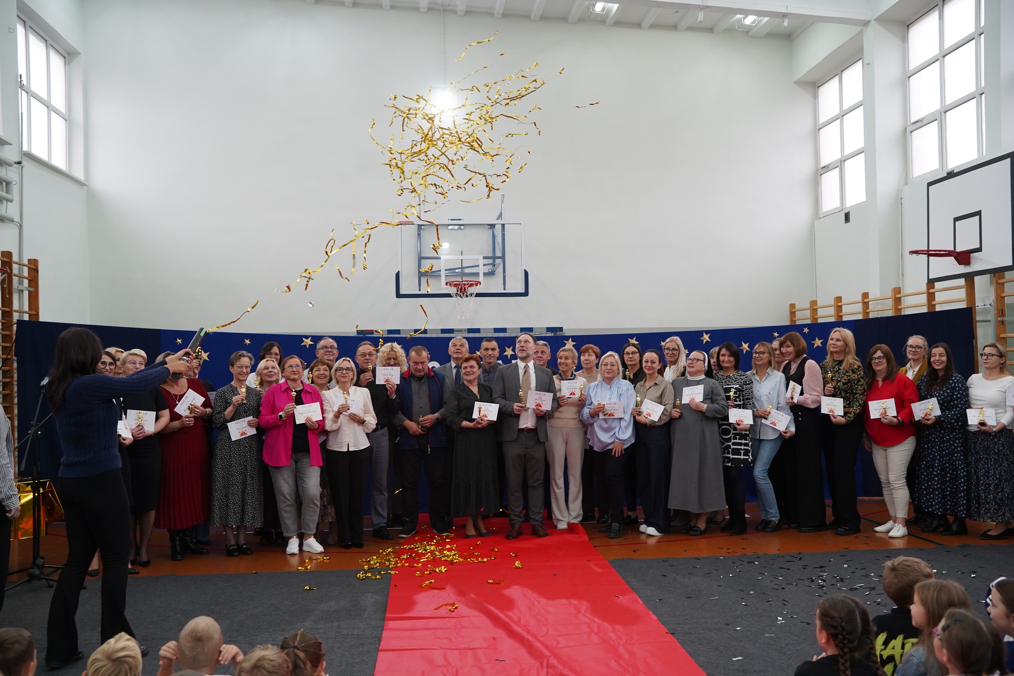 Na zdjęciu widać grupę osób stojących w sali gimnastycznej, trzymających dyplomy lub certyfikaty. Przed nimi leży czerwony dywan, a w powietrzu unosi się złote konfetti.