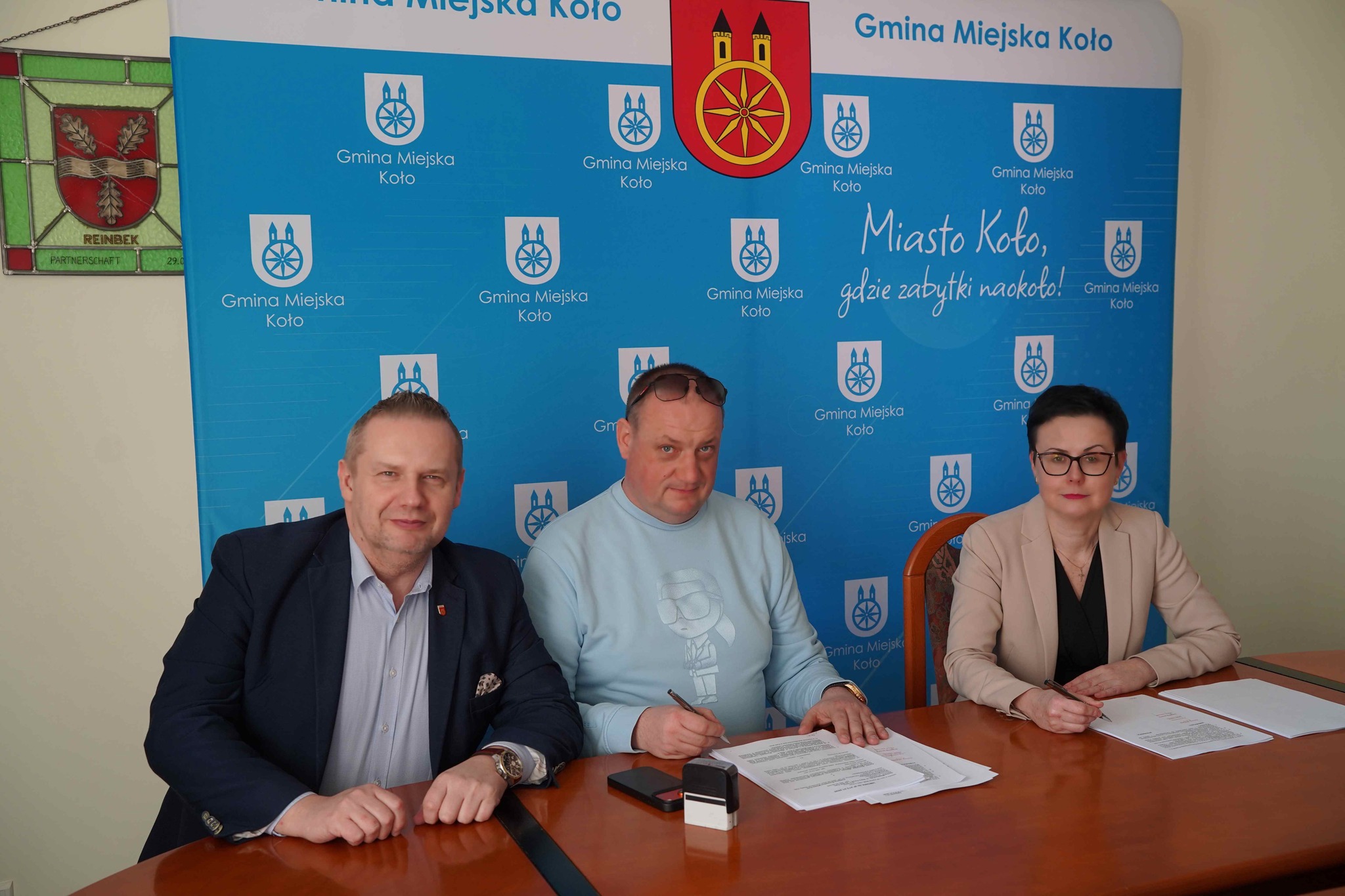 Na zdjęciu widać trzy osoby siedzące przy stole (po prawej kobieta, a po lewej mężczyźni) podpisujące umowę, W tle widać ściankę reklamową Gminy Miejskiej Koło.
