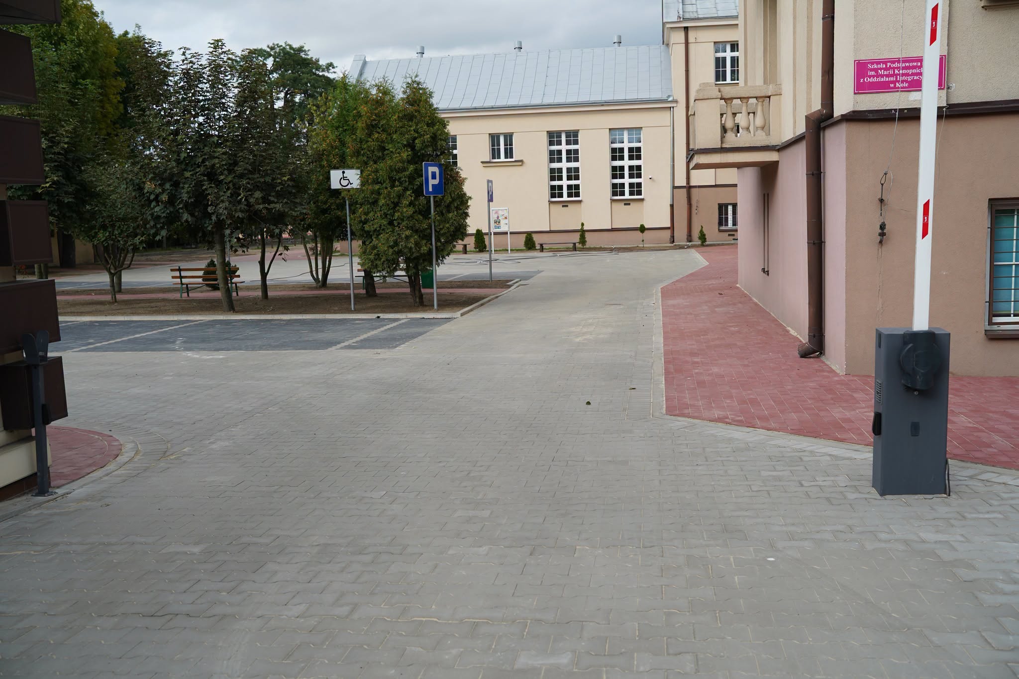Plac wyłożony szarą kostką brukową przy budynku szkoły. Po lewej wyznaczone miejsce postojowe z oznakowaniem dla osób z niepełnosprawnościami, znak P. Po prawej różowy chodnik, szary słupek szlabanu, maszt i tablica z nazwą szkoły podstawowej.