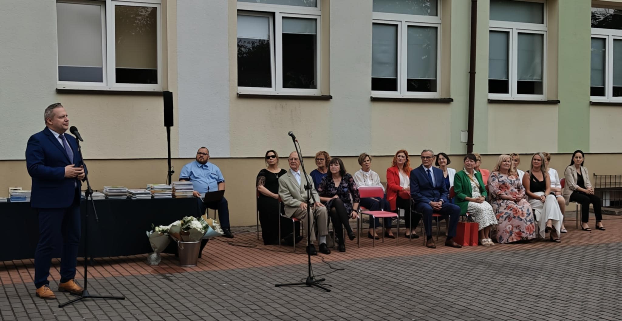 Na zdjęciu widać uroczystość szkolną na świeżym powietrzu – mężczyzna w garniturze przemawia do mikrofonu, a za nim siedzi grupa nauczycieli i gości. Obok stoją kwiaty i stosy książek, prawdopodobnie nagrody.