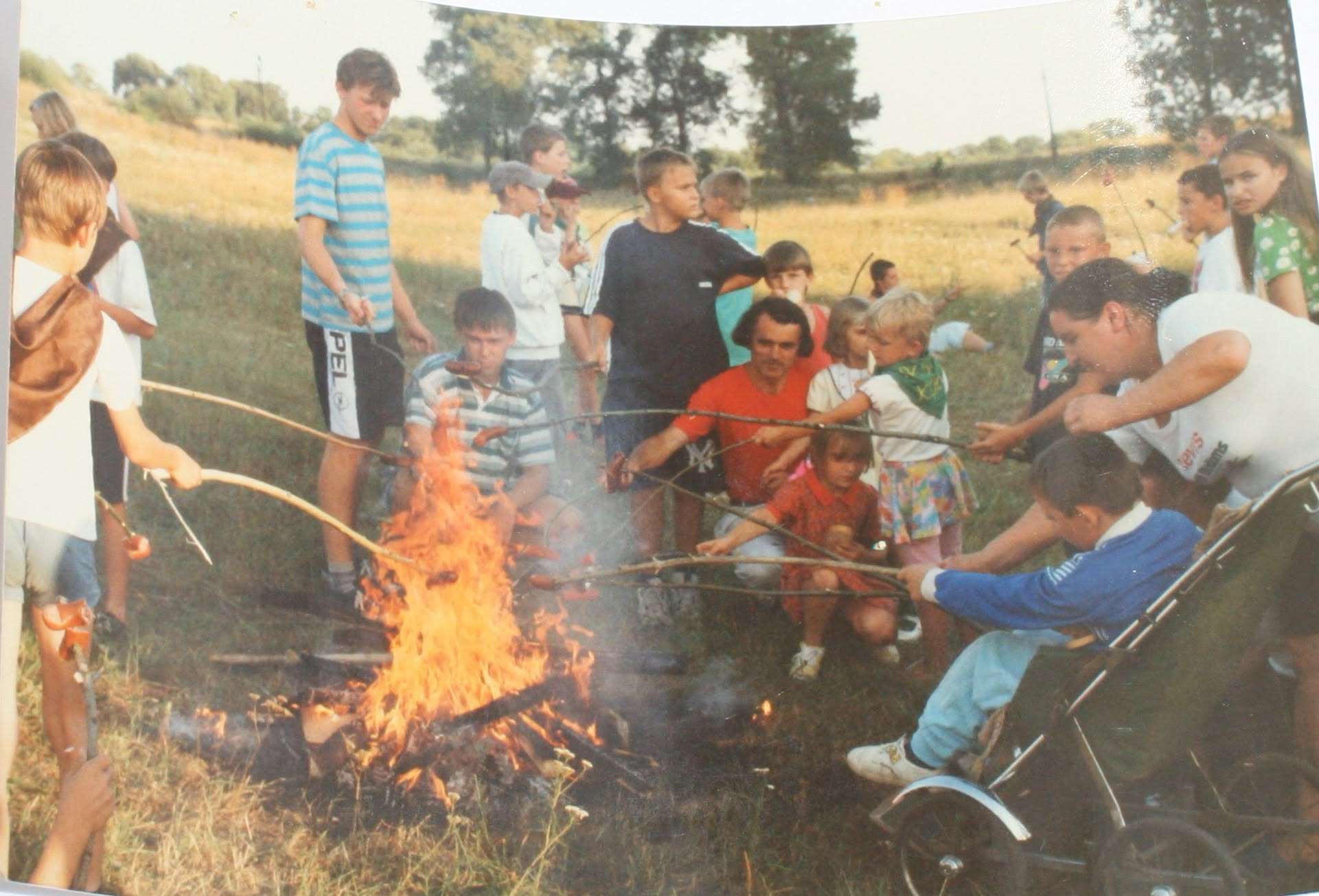 Grupa dzieci i dorosłych piecze kiełbaski na patyках nad ogniskiem rozpalonym na łące. W tle widoczne drzewa i pola uprawne. Przy ogniu siedzi niemowlę w wózku. Zdjęcie analogowe, prawdopodobnie z lat 90. XX wieku.