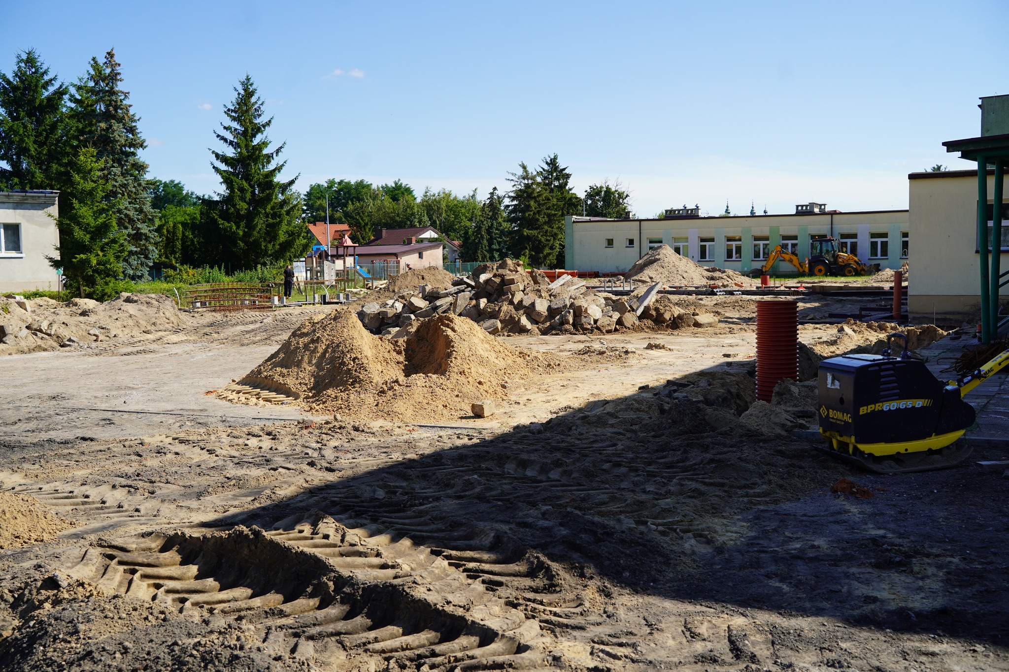 Zdjęcie przedstawia plac budowy z koparką, hałdami piasku i gruzu oraz widocznym budynkiem w tle, prawdopodobnie w trakcie rozbudowy lub modernizacji