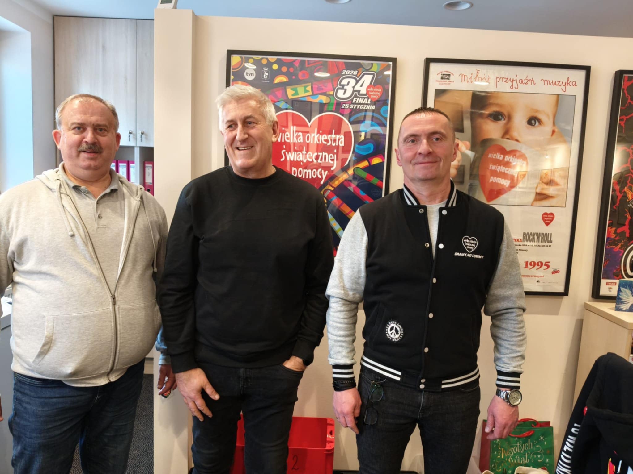 Na zdjęciu widać trzech stojących mężczyzn. Są to członkowie Kolskiego Sztabu WOŚP, a zdjęcie zrobiono w siedzibie fundacji WOŚP w Warszawie. Za nimi na ścianie wiszą plakaty z poprzednich finałów WOŚP.