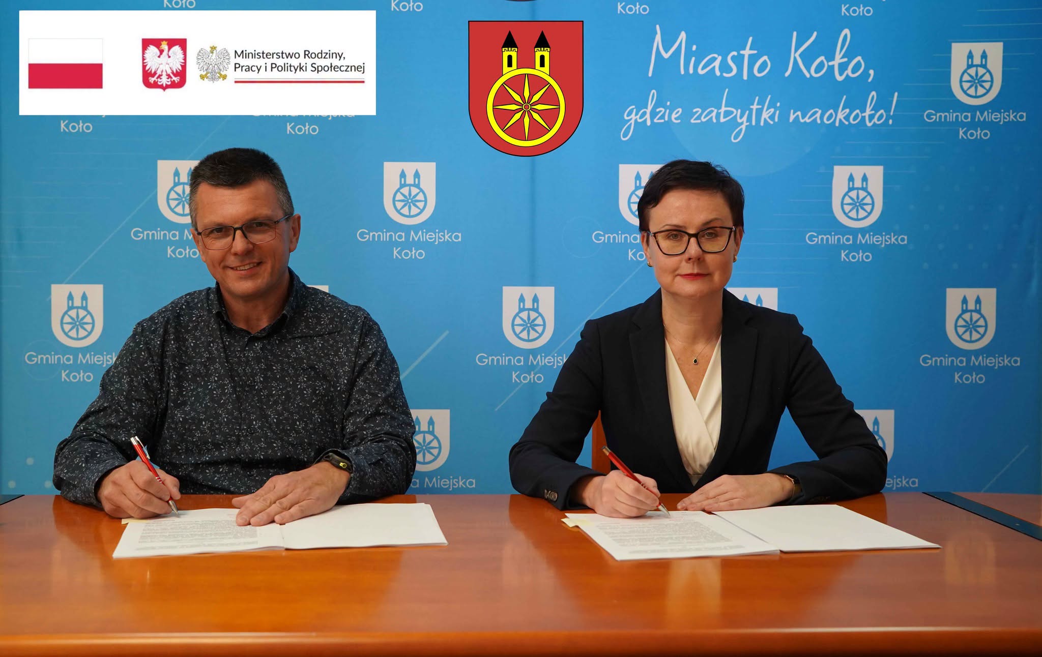 Dwie osoby siedzące przy stole (po prawej kobieta, a po lewej mężczyzna) podpisujące umowę. W tle widać ściankę reklamową Gminy Miejskiej Koło.