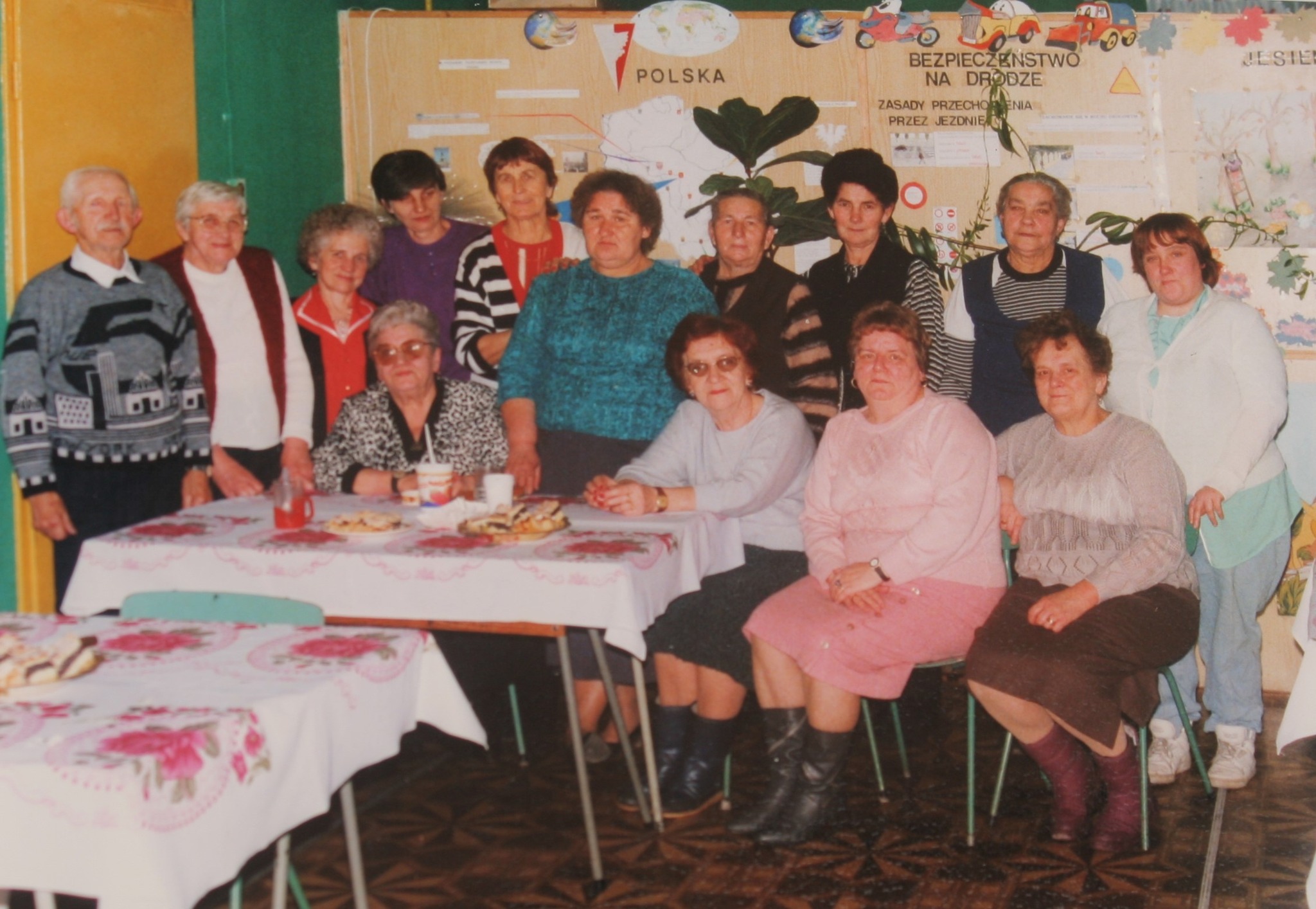 Grupa seniorów pozuje do zdjęcia w świetlicy klubowej. Część osób siedzi na krzesełkach przy stole, część stoi za osobami siedzącymi. W tle widać gazetkę ścienną. 