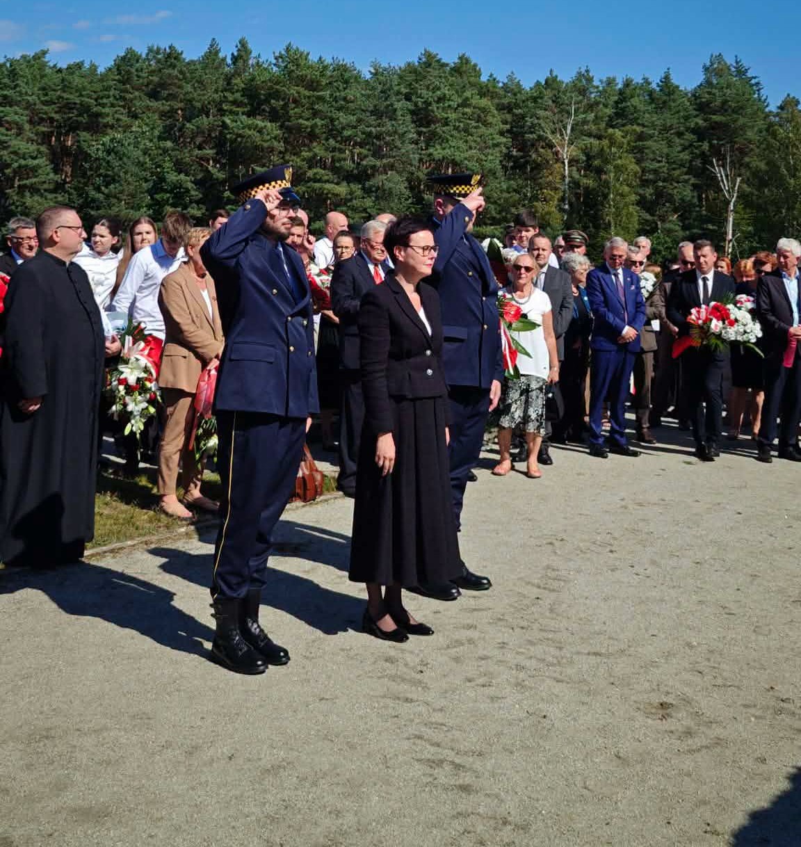 Uroczystość patriotyczna na piaszczystym placu przy lesie. Dwóch funkcjonariuszy w granatowych mundurach salutuje, obok kobieta w czarnym kostiumie stoi na baczność. W tle tłum uczestników z biało-czerwonymi wiązankami, słoneczny dzień.