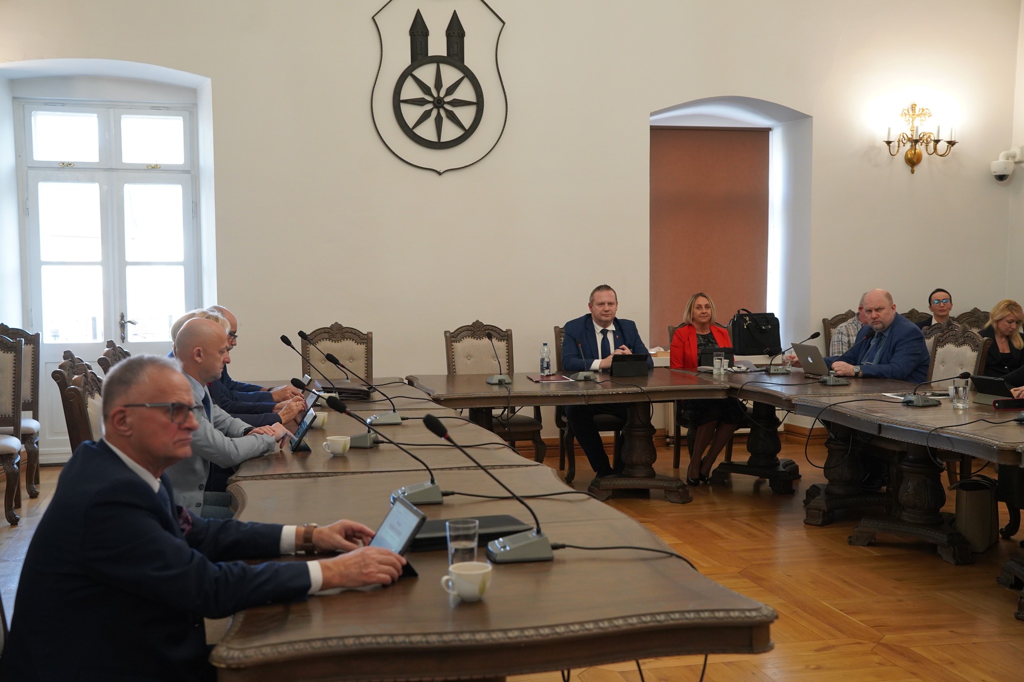 Na zdjęciu widać obrady sesji Rady Miejskiej Koła– przy stołach siedzą radni z tabletami oraz burmistrz wraz z Panią Skarbnik, na stołach stoją mikrofony, a na ścianie u góry widoczny jest herb miasta.
