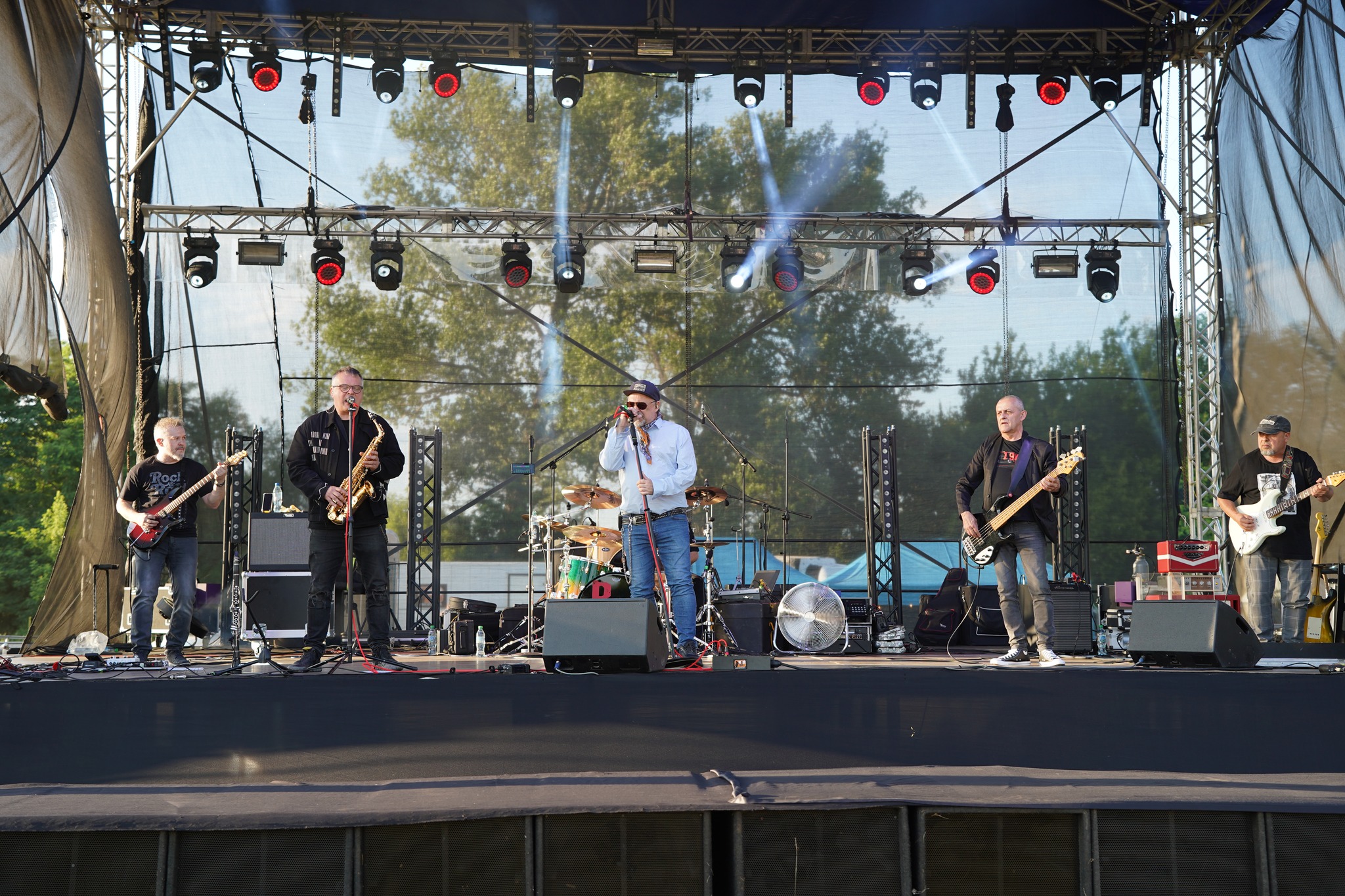 Na zdjęciu widać zespół muzyczny występujący na dużej plenerowej scenie. Artyści grają na gitarach, saksofonie i perkusji. W centrum wokalista śpiewa do mikrofonu. Scena oświetlona jest reflektorami, w tle widoczne są drzewa.