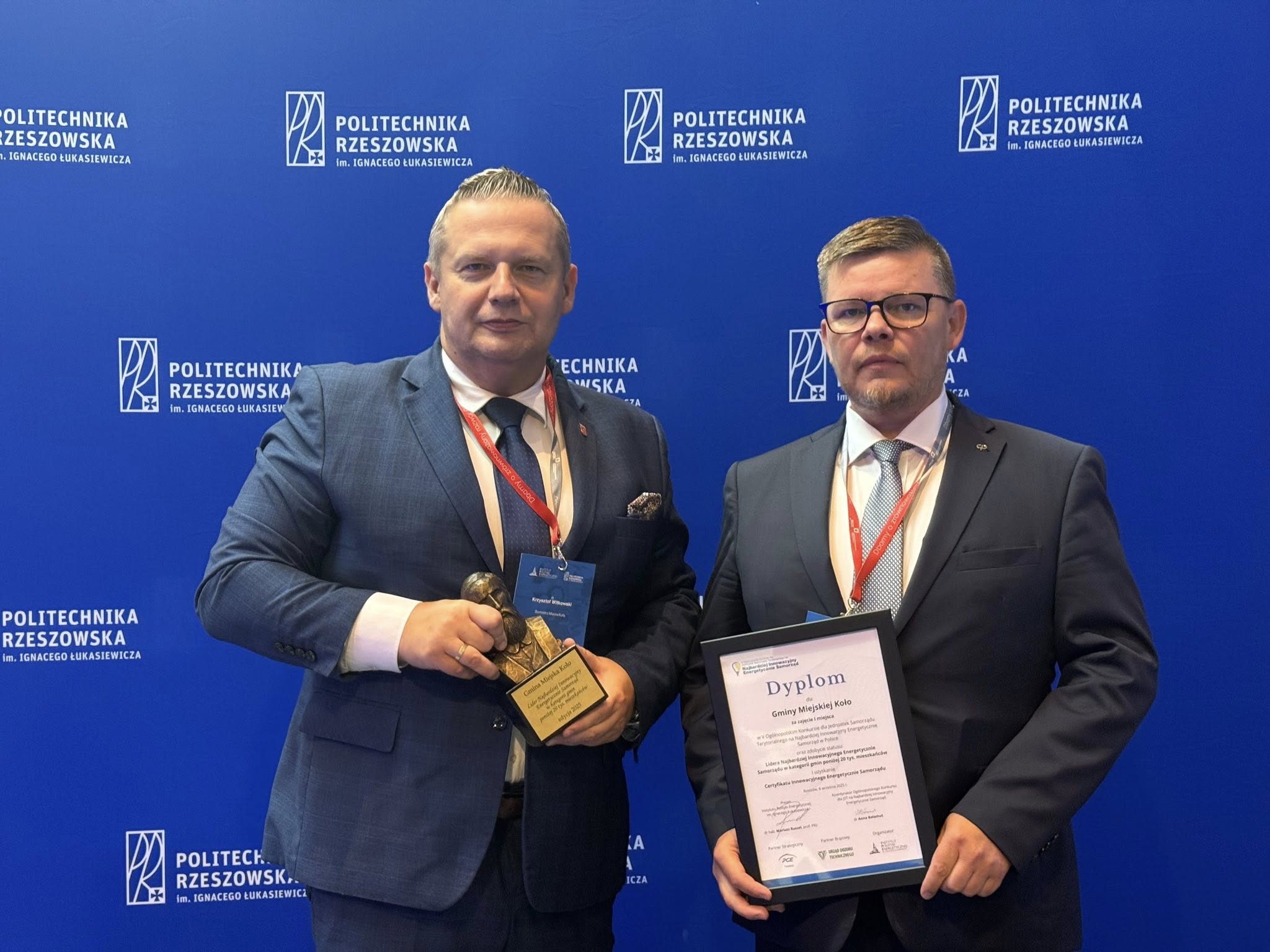 Na zdjęciu widać dwóch mężczyzn w garniturach stojących na tle ścianki reklamowej Politechniki Rzeszowskiej. Mężczyzna z lewej trzyma w dłoniach statuetkę, a z prawej pamiątkowy dyplom w drewnianej czarnej ramce.