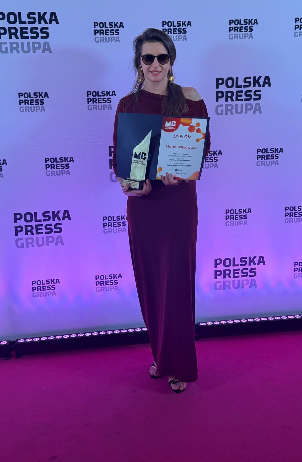 Kobieta w bordowej sukni stoi na tle ścianki Polska Press Grupy. Trzyma statuetkę oraz dyplom z napisem Marta Witkowska w konkursie Mistrzowie Budownictwa