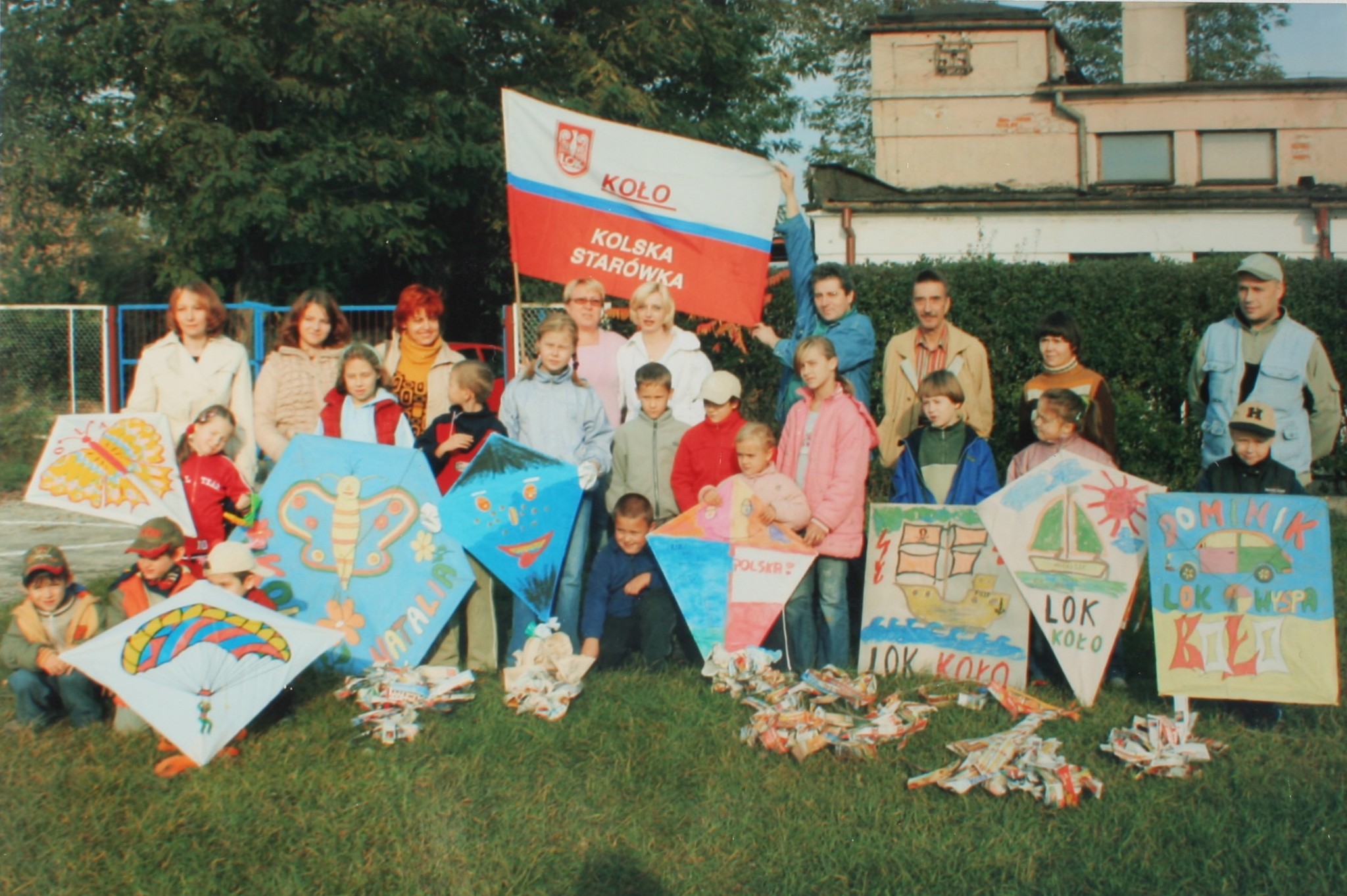 Grupa dzieci i dorosłych pozuje z kolorowymi latawcami i flagą z napisem Koło Kolska Starówka.