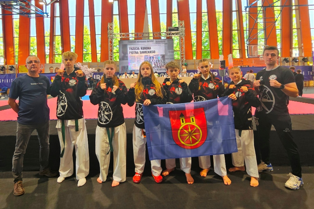 Grupa młodych zawodników sztuk walki w czarnych strojach klubowych pozuje z flagą miasta (herb z kołem i wieżą). Towarzyszą im dwaj trenerzy. W tle hala sportowa z banerem turnieju Trzecia Korona Piotra Sawickiego.
