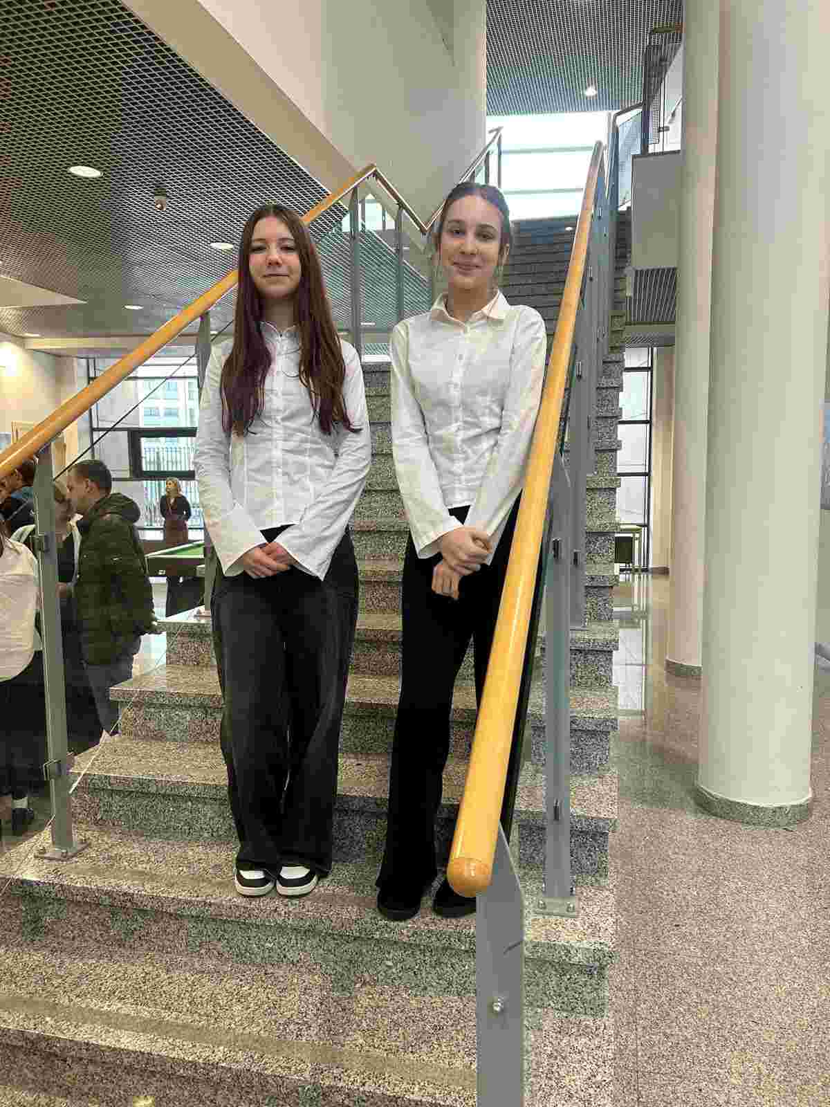 Na zdjęciu widać dwie dziewczyny pozujące do fotografii – stoją na schodach, ubrane w białe bluzki i stylowe, eleganckie spodnie. W tle, po lewej stronie, dostrzec można inne stojące osoby.