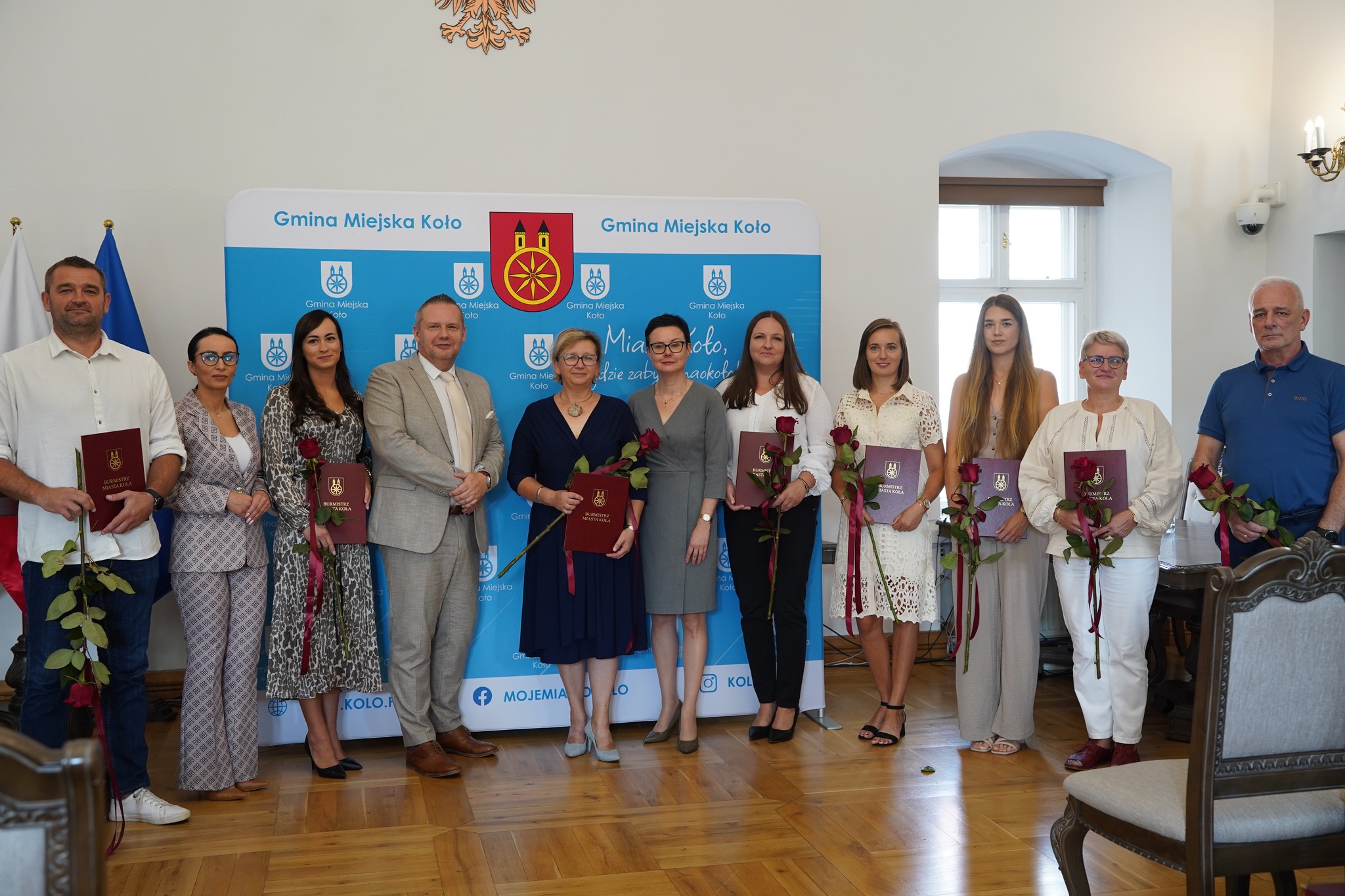 Na zdjęciu widać grupę osób stojących w sali urzędu, trzymających dyplomy i róże – to prawdopodobnie uroczystość wręczenia awansów w Gminie Miejskiej Koło