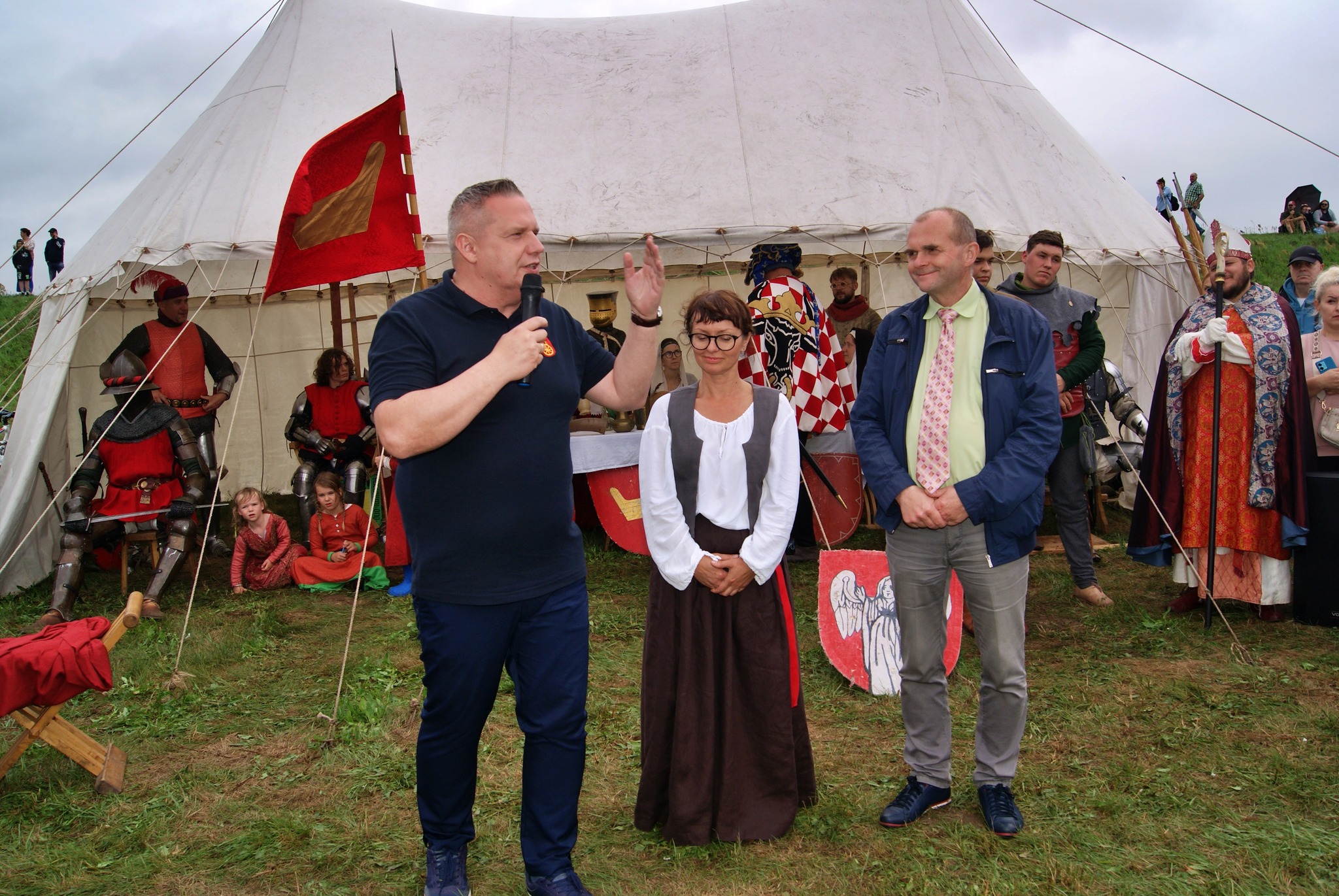 Na zdjęciu widać wydarzenie historyczne (festyn) – trzy osoby stoją przed namiotem, wokół znajdują się ludzie w strojach średniowiecznych, w tym rycerze w zbrojach.