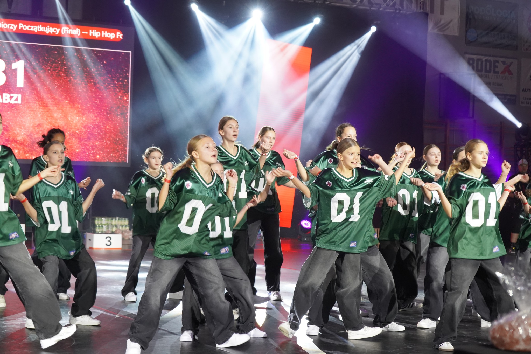 Na zdjęciu widać grupę młodych tancerek podczas występu na scenie, ubranych w zielone koszulki sportowe z numerem 01 i ciemne spodnie. Scena jest oświetlona kolorowymi reflektorami, a w tle widoczny jest ekran z napisem w języku polskim.