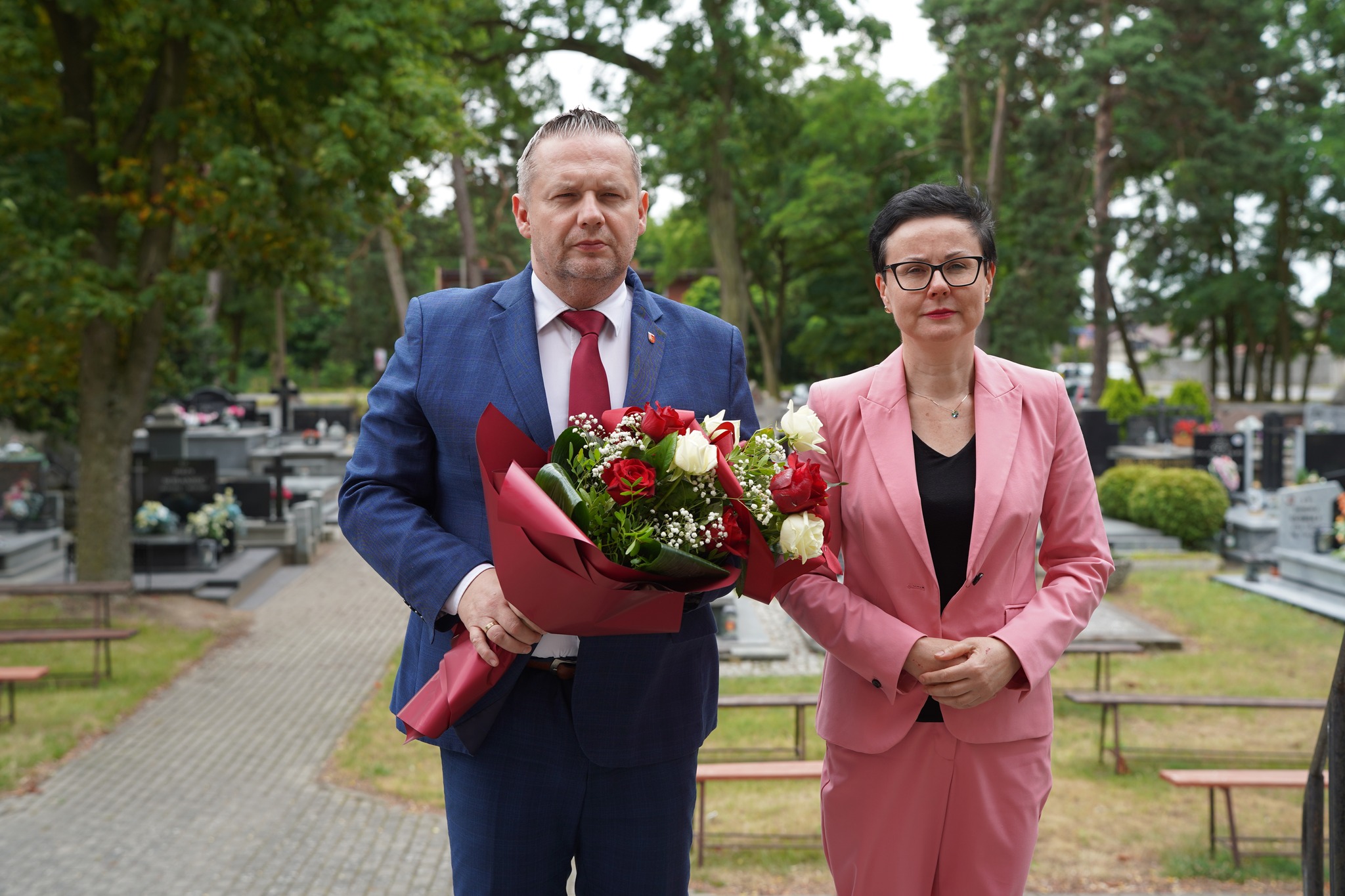 Na zdjęciu widoczni są mężczyzna i kobieta stojący na cmentarzu. Mężczyzna trzyma bukiet kwiatów, składają hołd Ofiarom Ludobójstwa na Wołyniu.