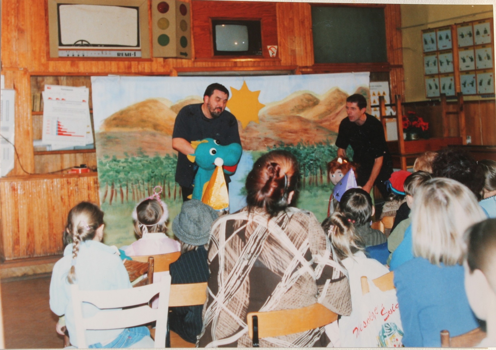Na zdjęciu widać przedstawienie dla dzieci: dwóch mężczyzn prowadzi teatrzyk z pacynkami przed malowaną scenografią, a przed nimi siedzi grupa dzieci i dorosłych oglądających występ w sali.