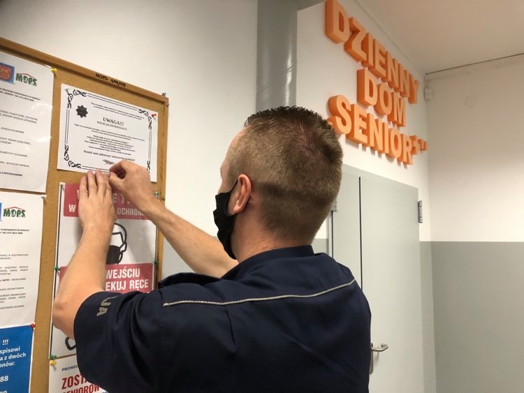 Policjant wiesza ulotkę informacyjną na tablicy ogłoszeniowej w Dziennym Domu Seniora.