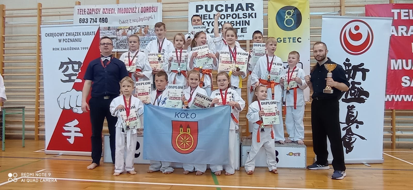 Grupowe zdjęcie kolskich zawodniczek i zawodników z dyplomami i pucharami wraz z opiekunami.