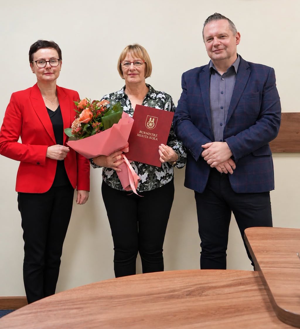 Trzy osoby pozują do zdjęcia w urzędzie. Kobieta w środku trzyma bukiet pomarańczowych róż i czerwoną teczkę z napisem Burmistrz Miasta Koła. Obok stoją kobieta w czerwonej marynarce i mężczyzna w granatowej marynarce.