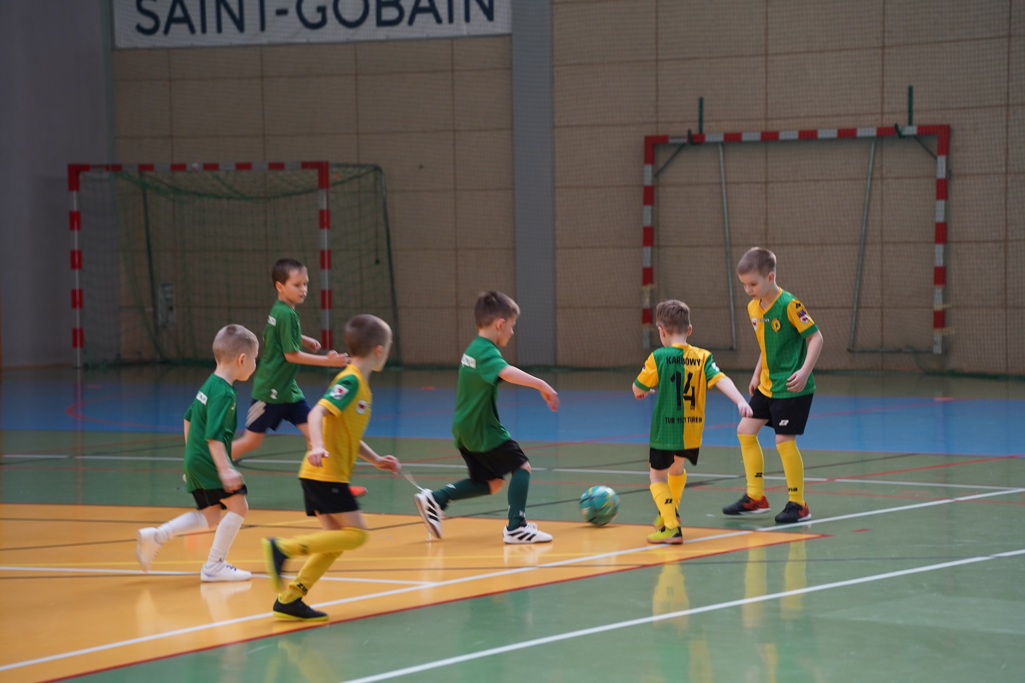 Na zdjęciu widać sześciu chłopców podczas emocjonującego meczu piłkarskiego na hali sportowej — w tle stoją bramki, a zawodnicy w zielonych oraz żółto-zielonych koszulkach walczą o piłkę na boisku.