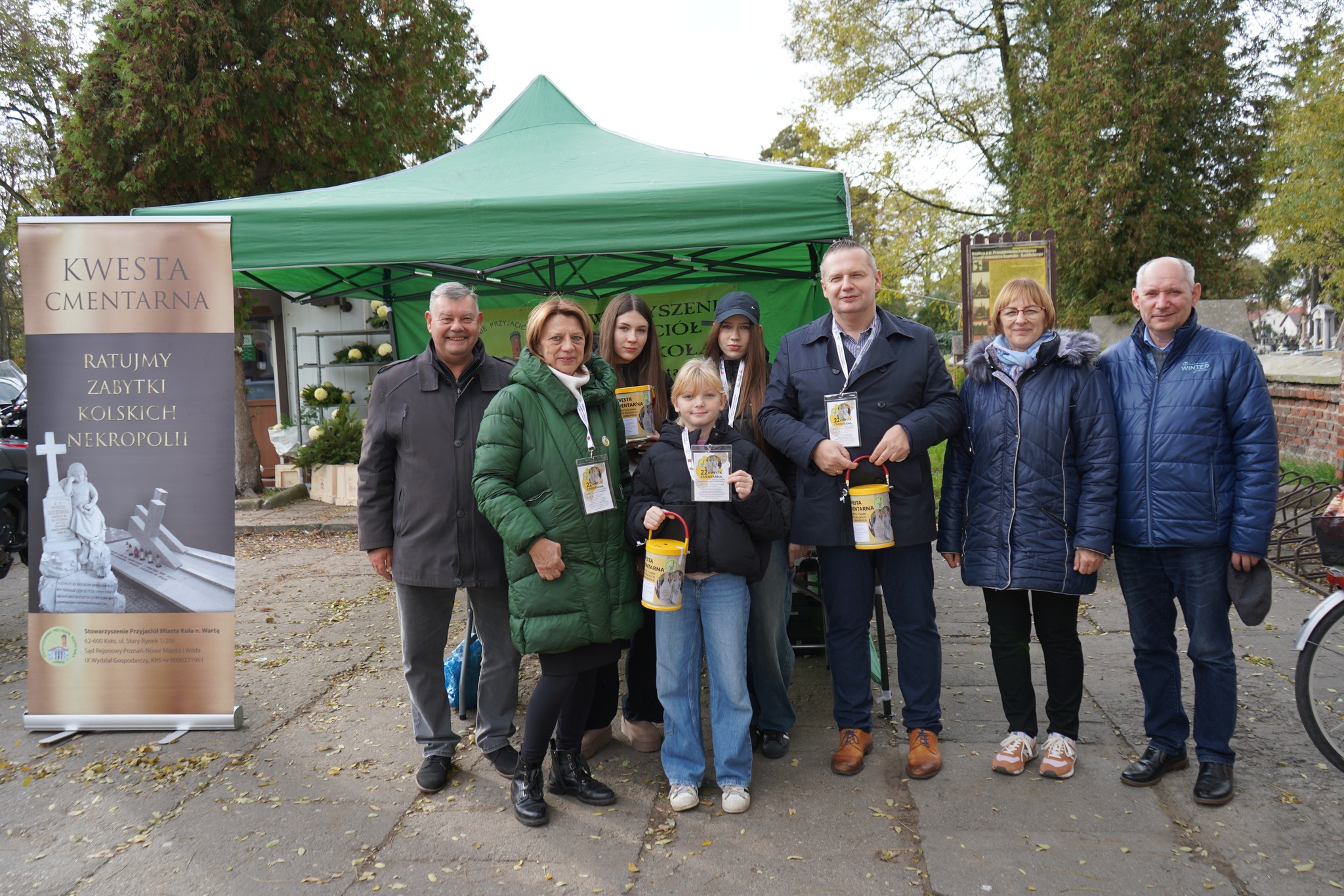 Na zdjęciu grupa osób stojących przed zielonym namiotem przed cmentarzem. Są uczestnikami kwesty na rzecz ratowania zabytków nekropolii w mieście Koło. Kilka osób trzyma puszki kwestarskie, w tle widać drzewa oraz cmentarne mury
