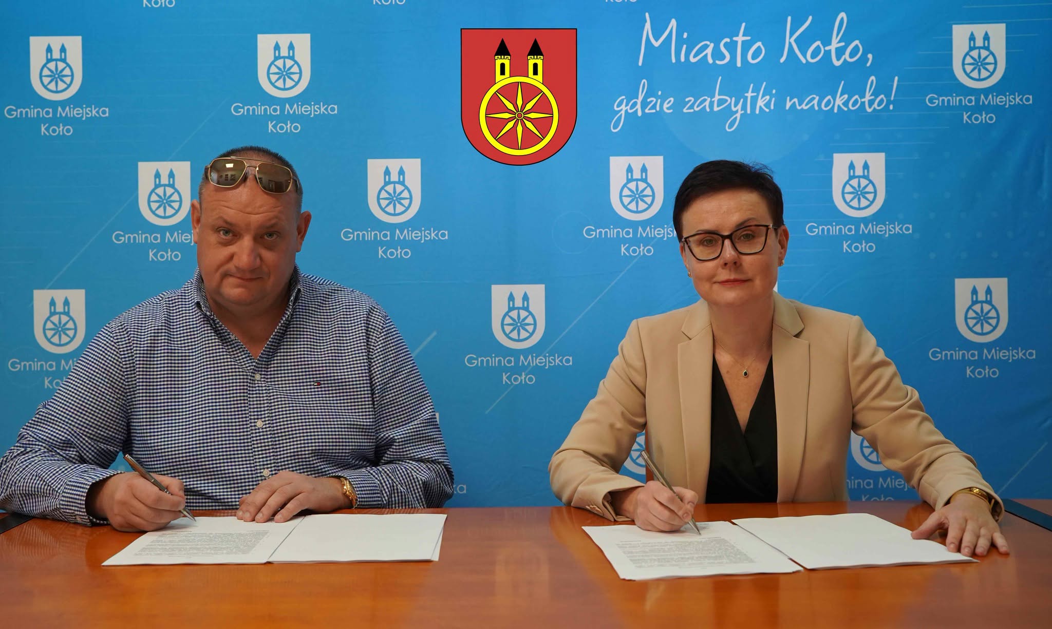 Dwie osoby siedzące przy stole (po prawej kobieta, a po lewej mężczyzna) podpisujące umowę, W tle widać ściankę reklamową Gminy Miejskiej Koło.