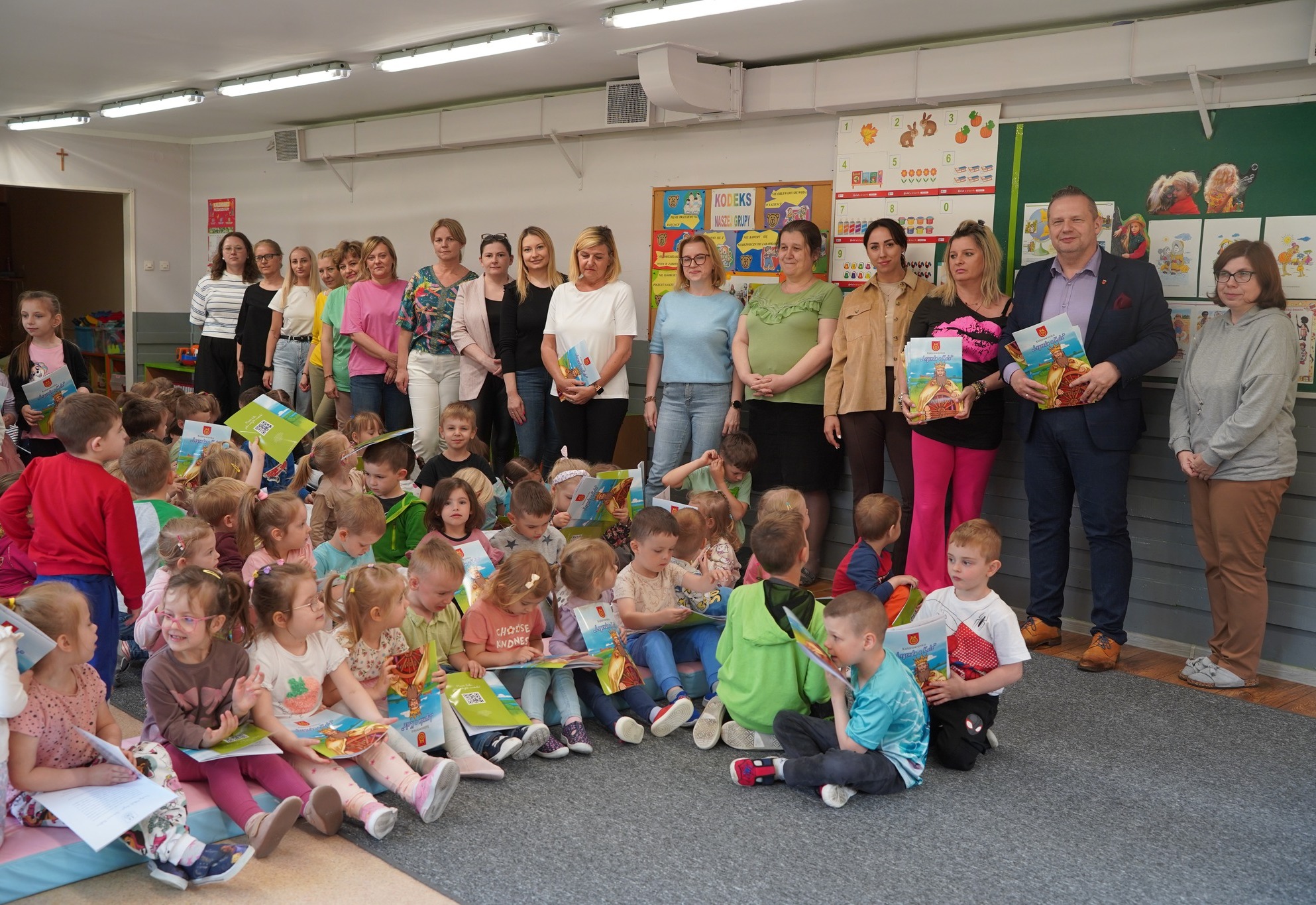 Zdjęcie przedstawia grupę dzieci siedzących na podłodze w przedszkolu oraz stojących za nimi dorosłych — prawdopodobnie nauczycieli i gości. Dzieci trzymają kolorowe książeczki, a atmosfera jest radosna i edukacyjna.