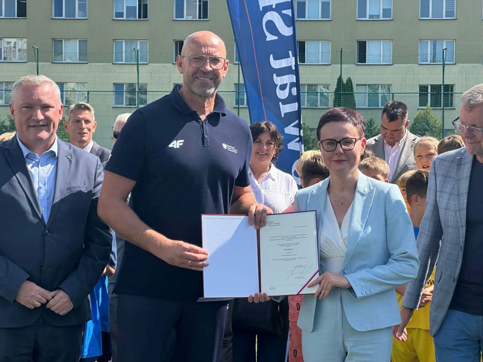 Mężczyzna w granatowej koszulce polo z logo 4F i kobieta w jasnobłękitnym żakiecie trzymają razem otwartą teczkę z dokumentem – promesą Ministerstwa Sportu i Turystyki. Wokół stoją urzędnicy i dzieci; w tle boisko z siatką, flaga i budynek.