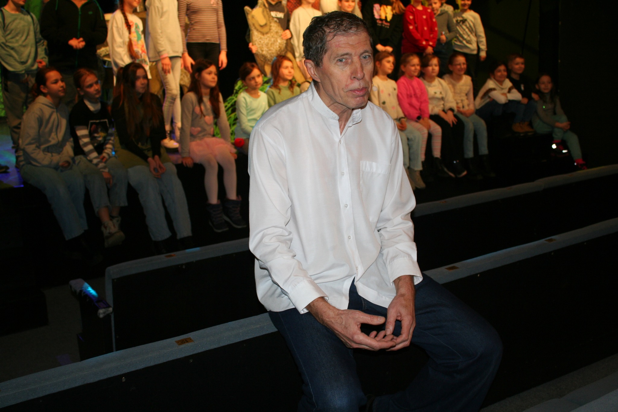 Na zdjęciu widać siedzącego mężczyznę w białej koszuli przed sceną w teatrze, w tle widać siedzącą na widowni młodzież.