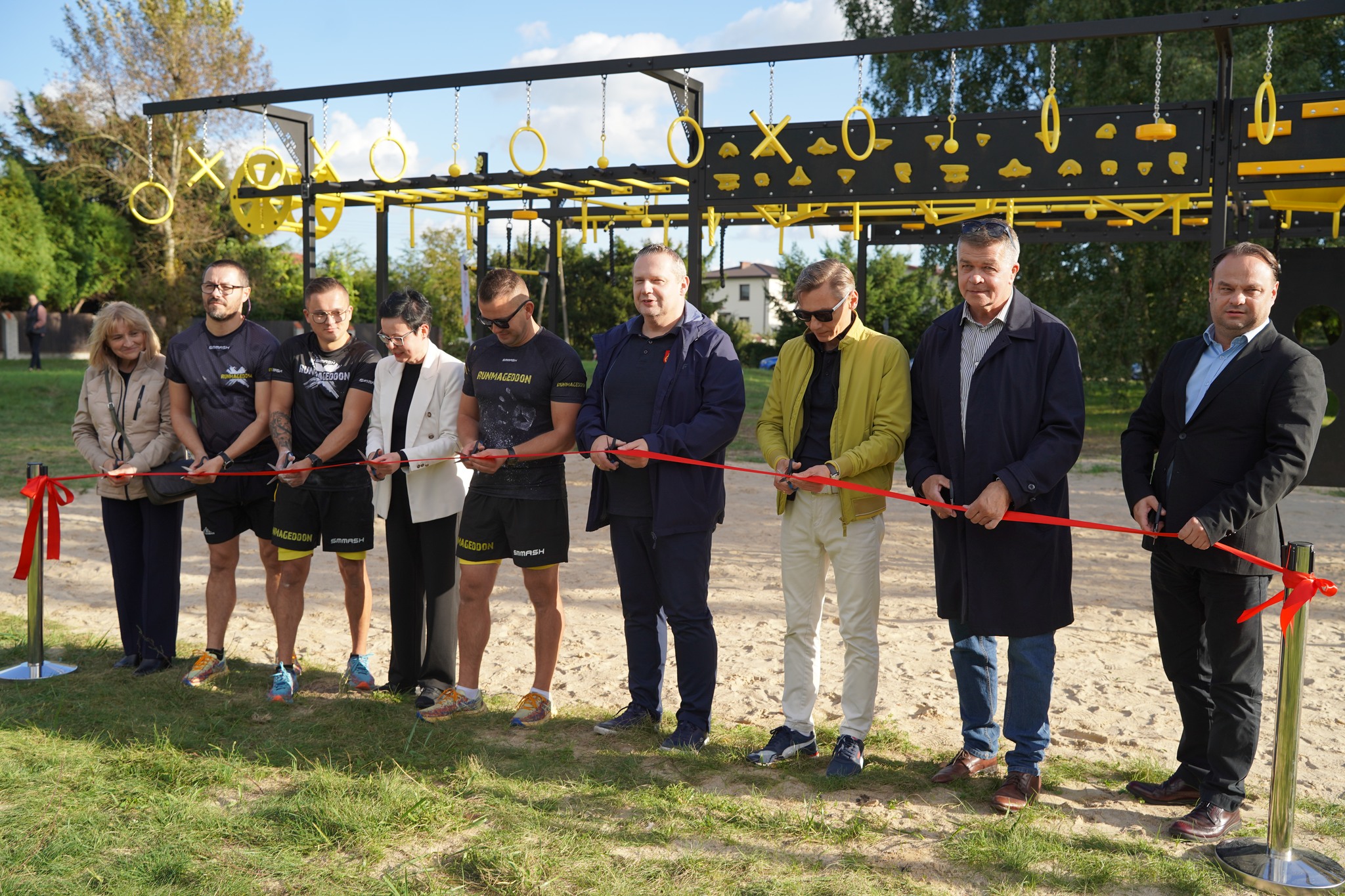 Na zdjęciu widać grupę osób biorących udział w uroczystym otwarciu toru sprawnościowego NINJA PARK w Kole. Uczestnicy przecinają wstęgę. W tle widoczny jest tor sprawnościowy oraz zielone drzewa.