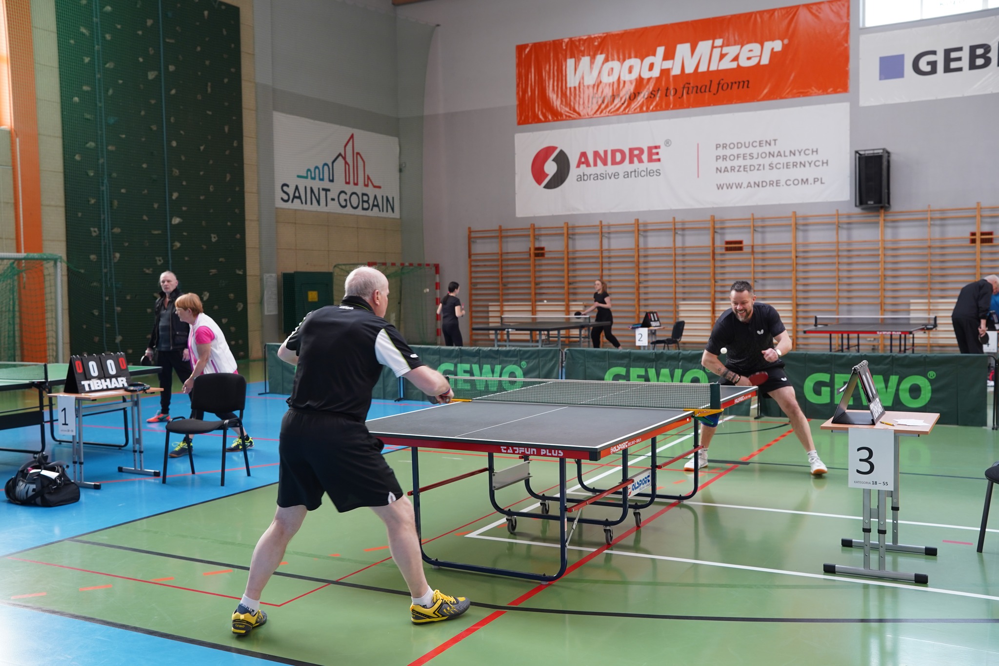 Na zdjęciu na pierwszym planie widać zawodników grających w tenisa stołowego w hali sportowej. W tle widać drabinki do ćwiczeń, wiszące banery reklamowe lokalnych firm oraz innych zawodników grających również w tenisa stołowego.