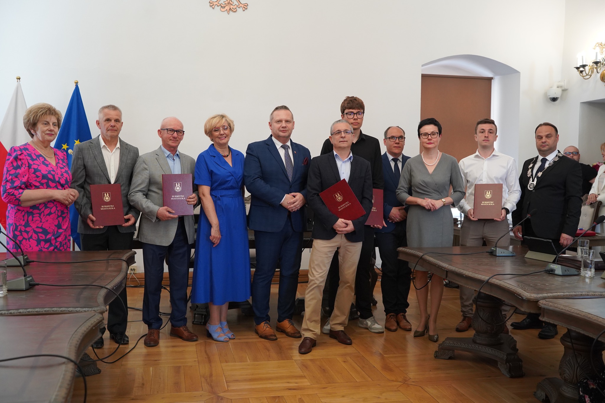 Na zdjęciu grupa osób pozujących do wspólnej fotografii w eleganckim wnętrzu urzędowym. Kilkoro z nich trzyma teczki z herbem miasta i napisem Burmistrz Miasta Koło, co sugeruje wręczenie nagród. W tle widoczne są flagi Polski i UE.