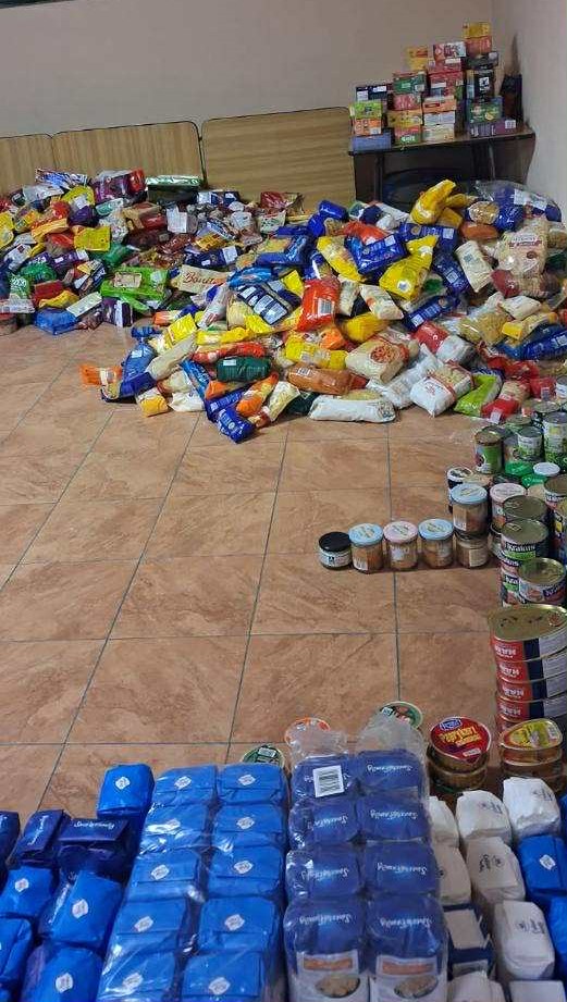 Na zdjęciu widać ustawione na podłodze w pomieszczeniu różnorodne produkty spożywcze — m.in. cukier, mąkę, konserwy, słodycze i słoiki — stanowiące zebrane dary w ramach zbiórki żywności.