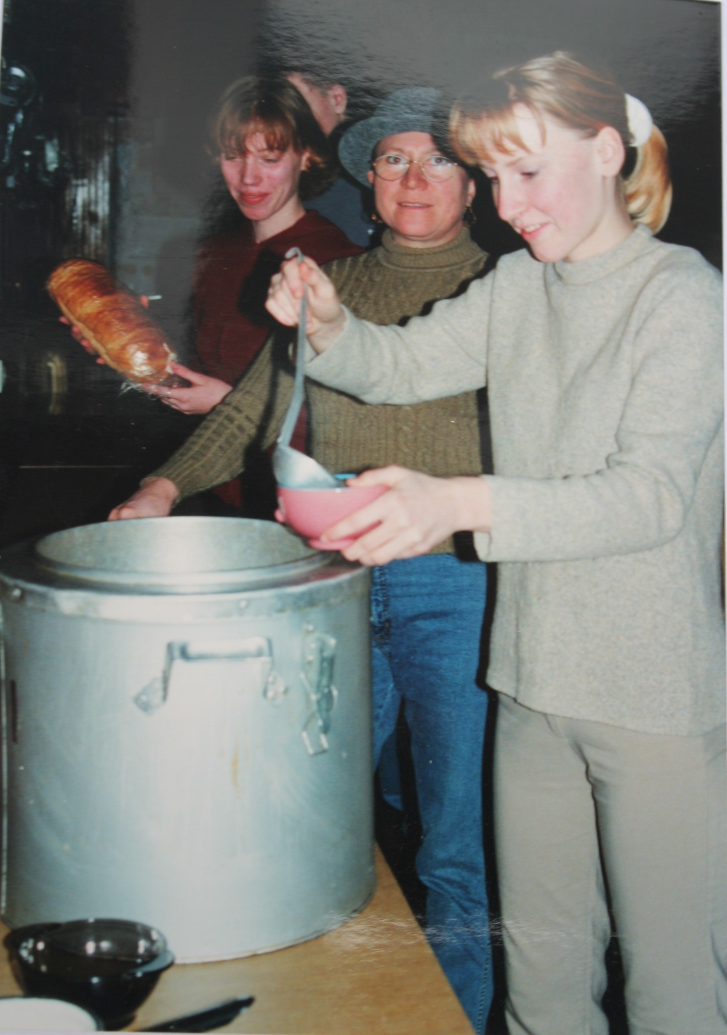 Na zdjęciu widać trzy kobiety — jedna nalewa wazówką z dużego kotła zupę do talerza, druga trzyma w dłoni bochenek chleba, a w tle stoi mężczyzna.