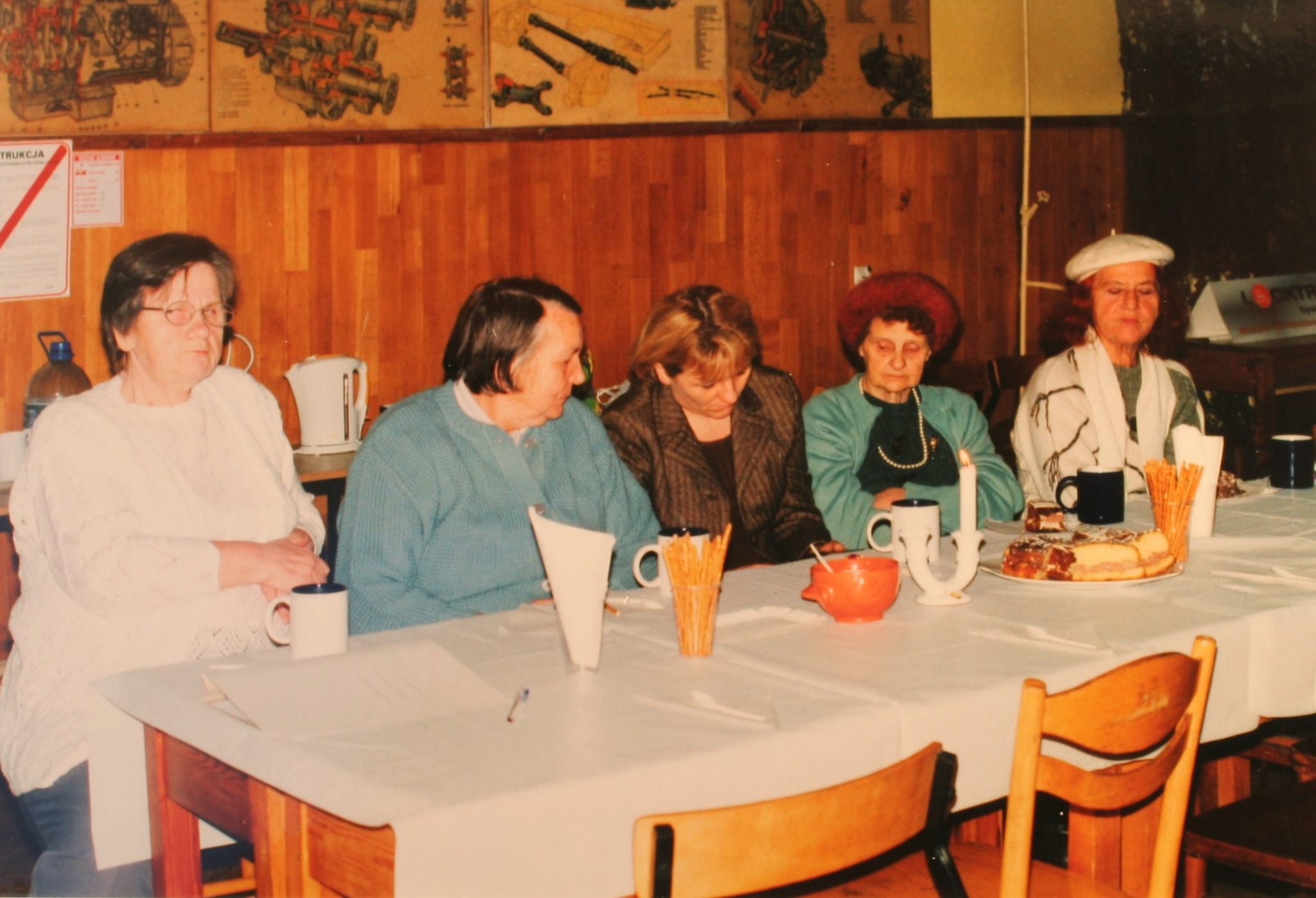 Na zdjęciu widać kilka starszych kobiet siedzących przy długim stole w świetlicy klubowej. Przed nimi stoją kubki, talerze z ciastem, świeca oraz drobne przekąski, co sugeruje kameralne spotkanie przy herbacie. Wnętrze ma drewniane ściany.