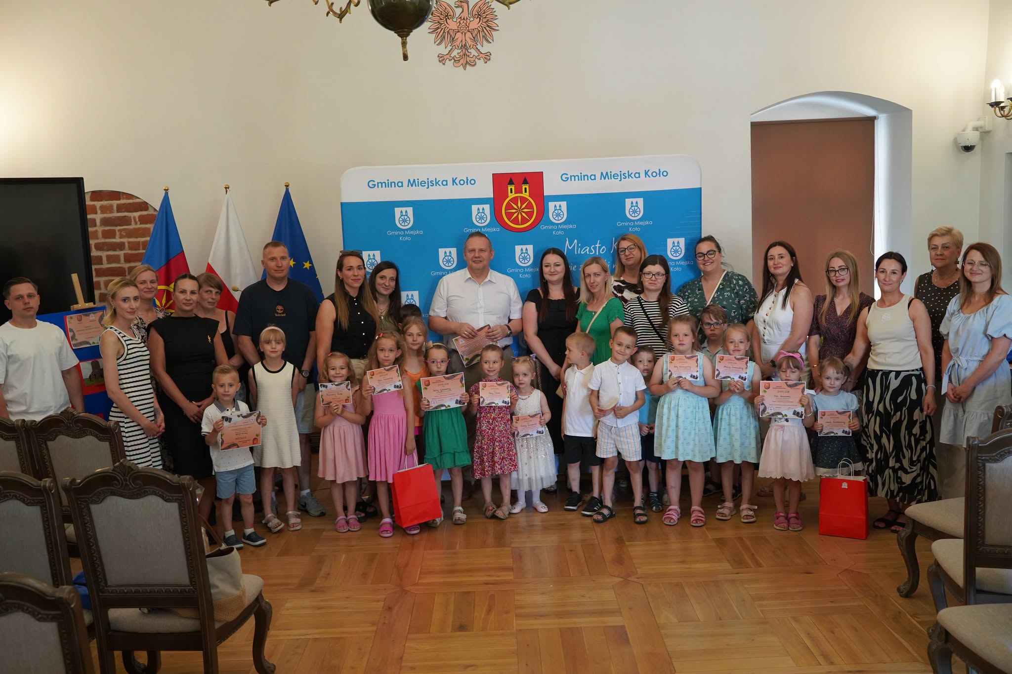 Grupowe zdjęcie dorosłych i małych dzieci trzymających dyplomy w sali z parkietem. W tle baner Gmina Miejska Koło oraz flagi Polski i UE. Mężczyzna w białej koszuli stoi pośrodku grupy.