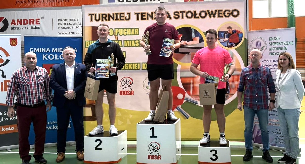 Zdjęcie przedstawia dekorację zwycięzców turnieju tenisa stołowego o Puchar Burmistrza Miasta Koła. Na podium widać trzech mężczyzn, trzymających w dłoniach puchary, dyplomy i nagrody. Obok nich stoją organizatorzy, w tle roll-upy organizatorów.